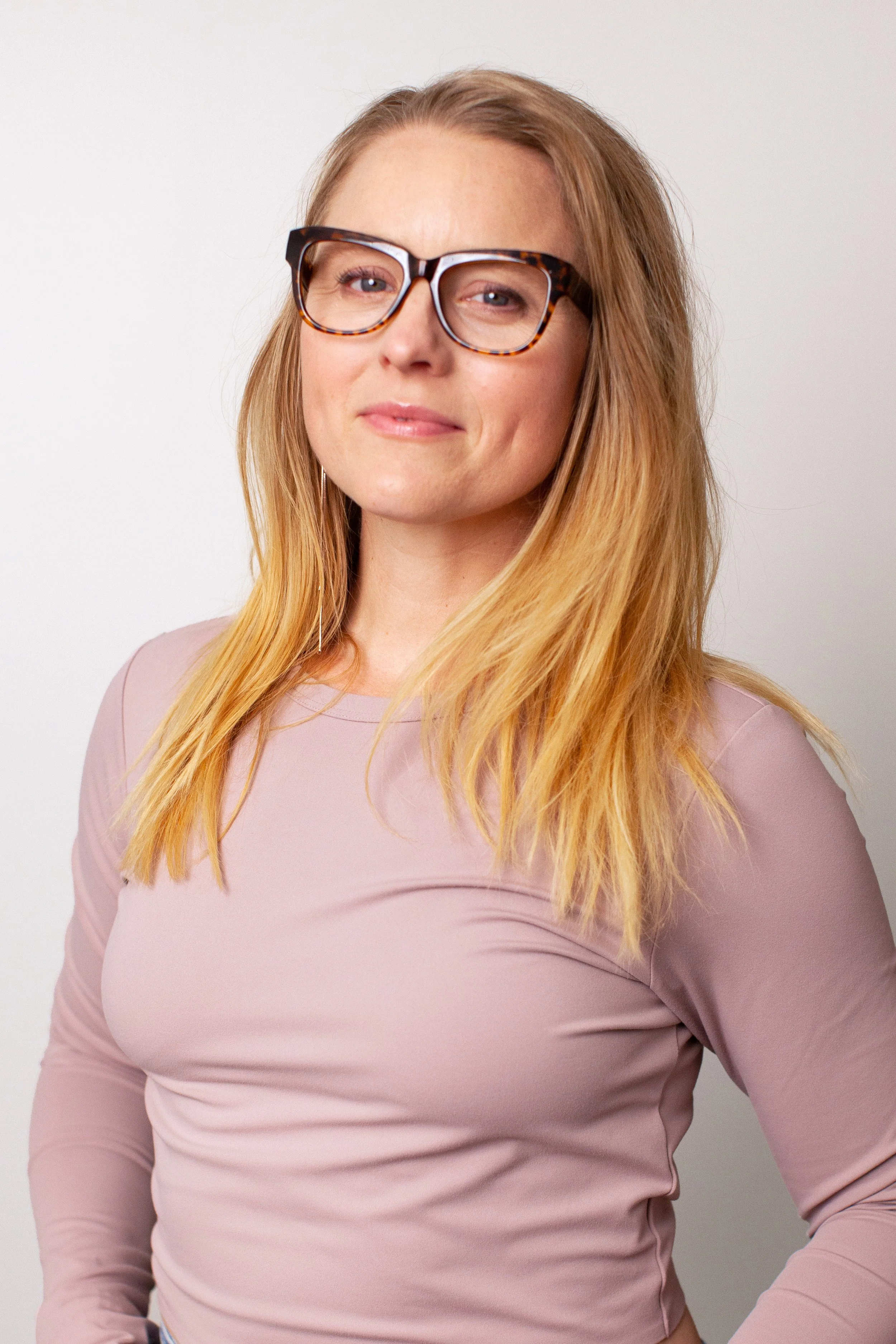 A woman with long red hair wearing tortoiseshell glasses and a mauve long-sleeve top, standing against a white background, looking at the camera with a slight smile.