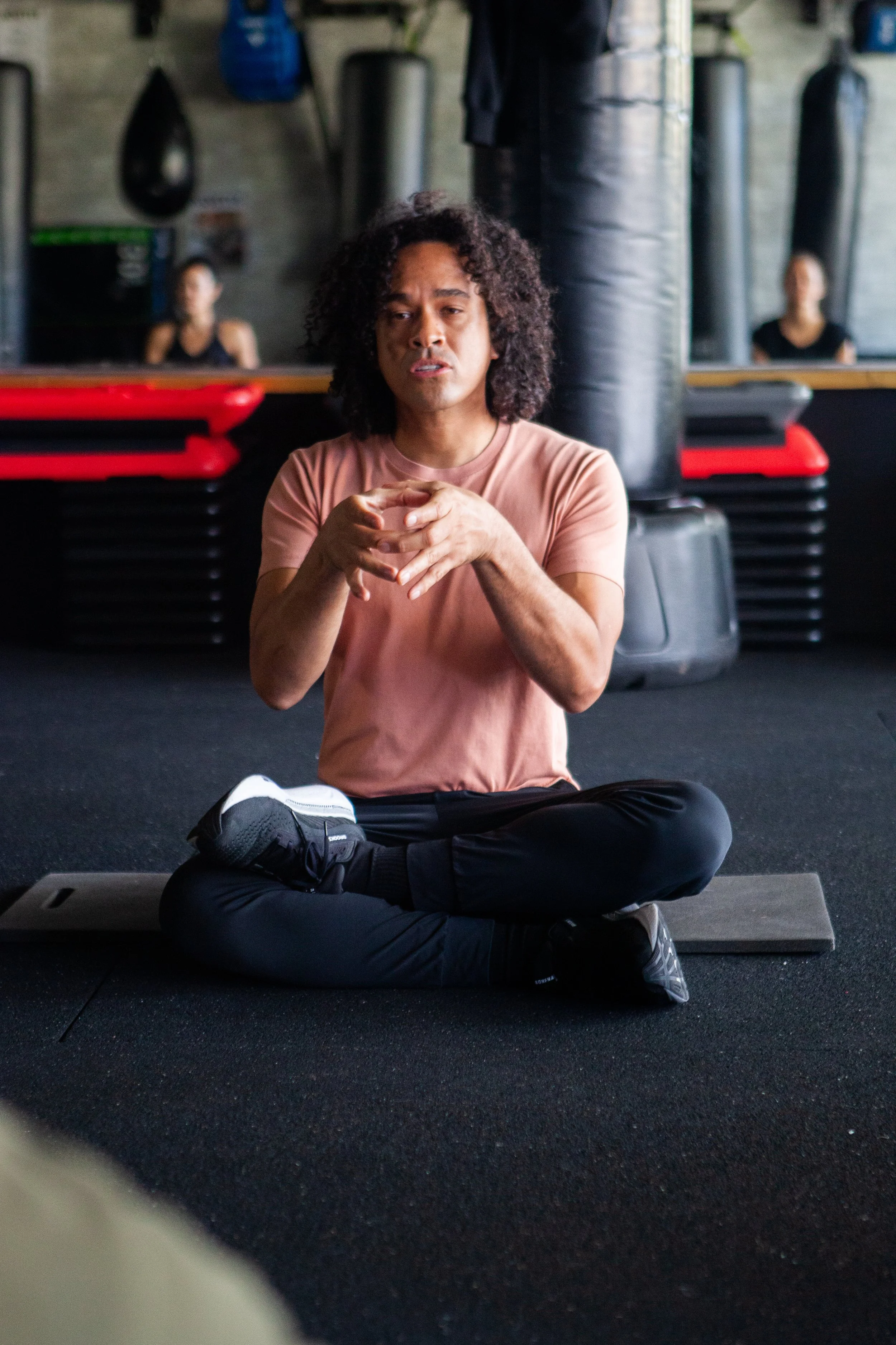 A man in workout clothes sitting cross-legged on the gym floor, speaking or explaining, with boxing equipment and mirrors in the background.