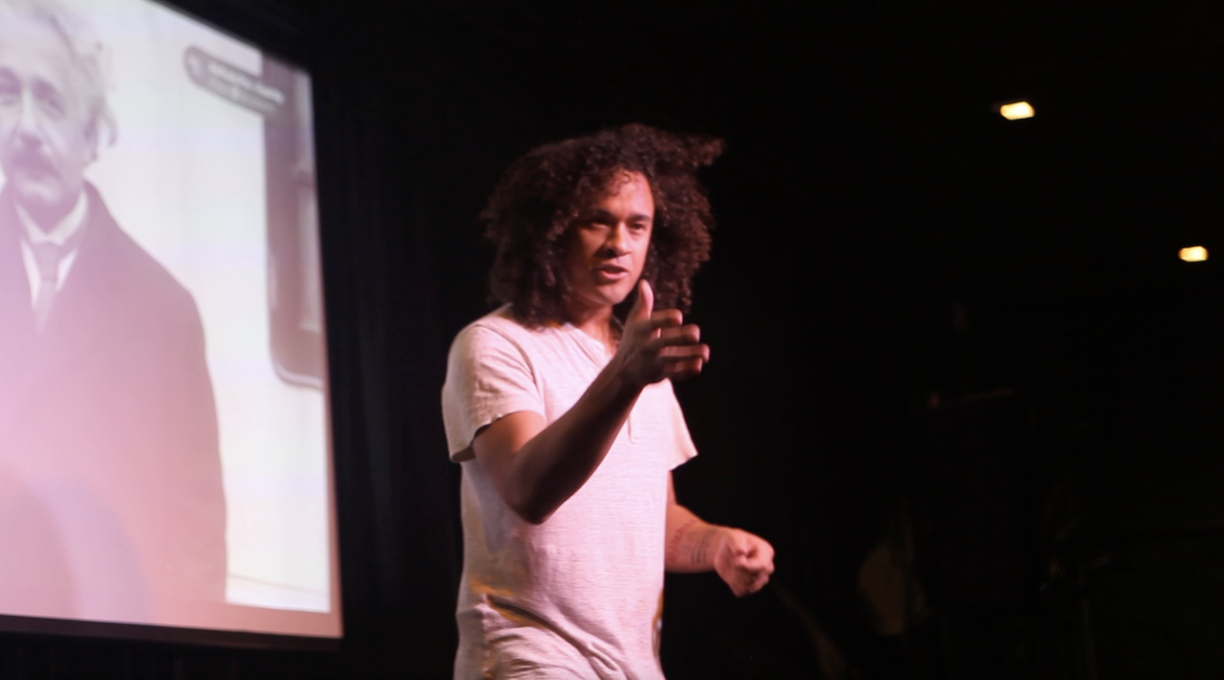 A person with curly hair speaking on stage, gesturing with their hand, in front of a projection screen showing an image of a man.