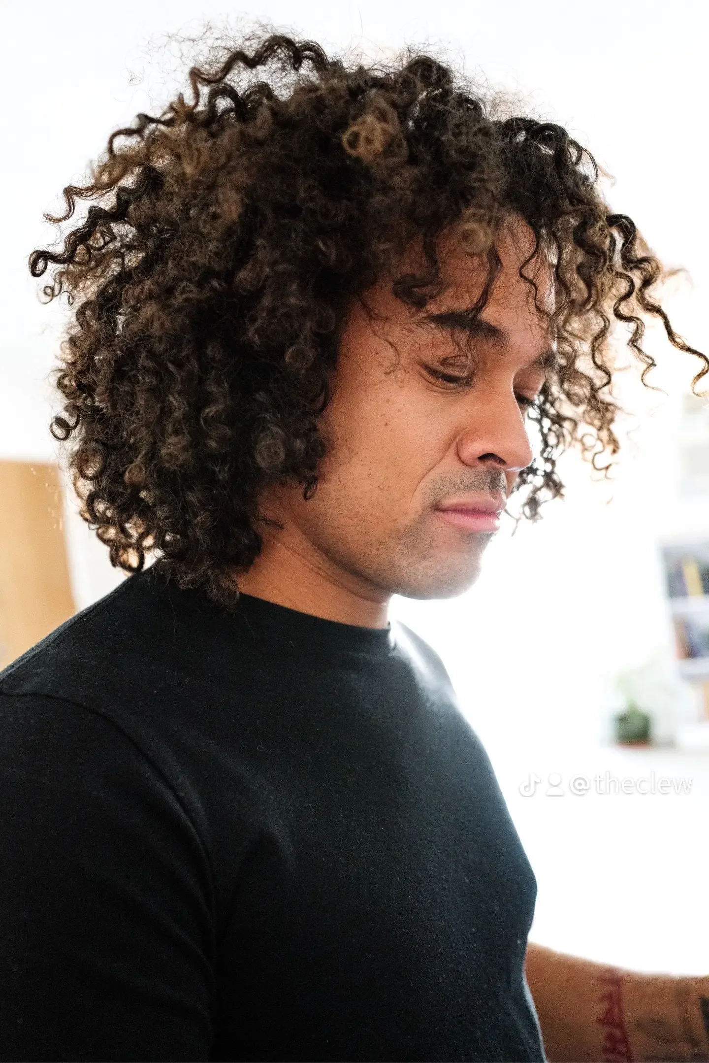 A man with curly dark hair, wearing a black shirt, looking down in an indoor setting.