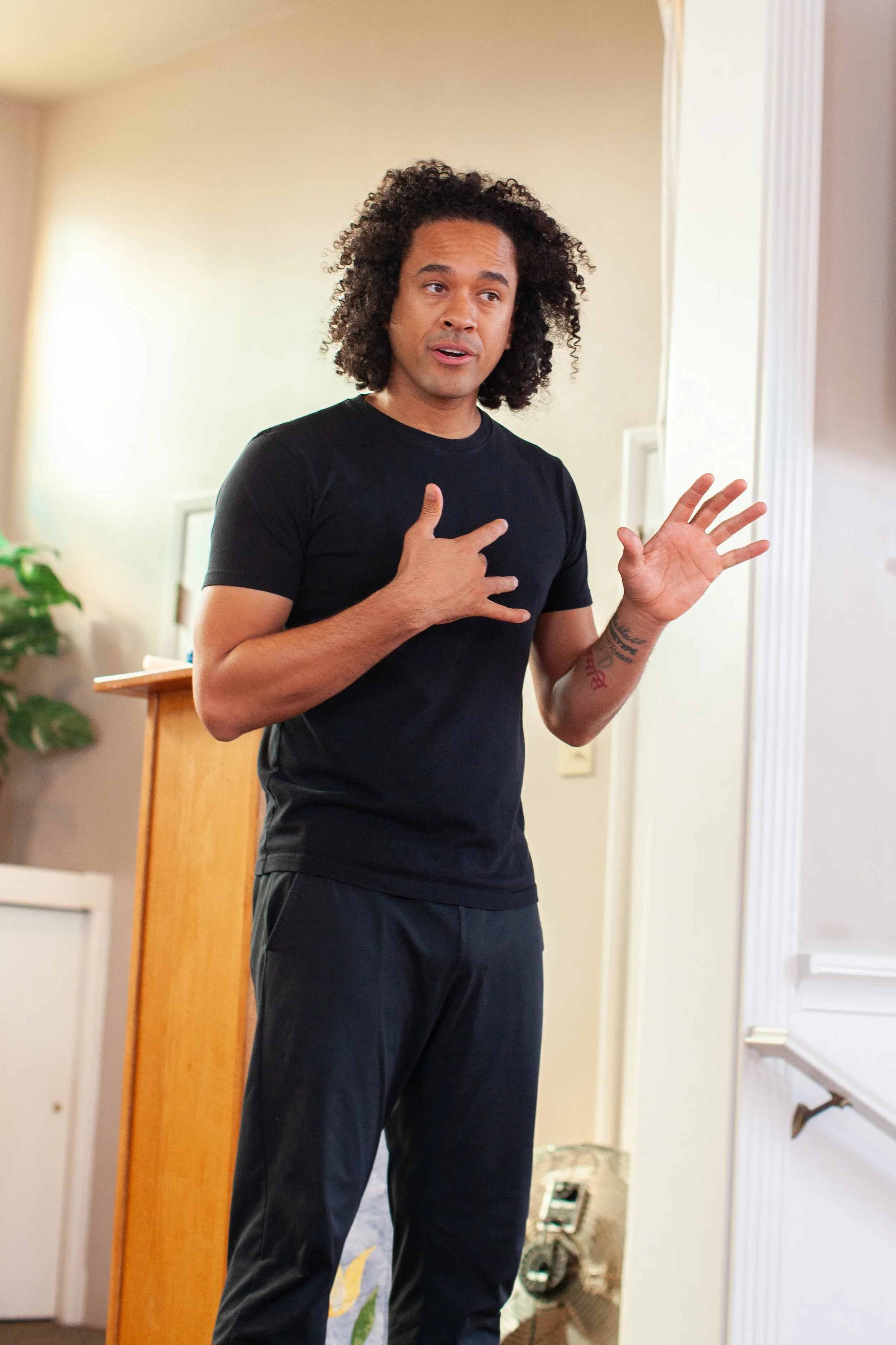 A man with curly hair, wearing a black t-shirt and black pants, is gesturing with his left hand and touching his chest with his right hand while speaking indoors.