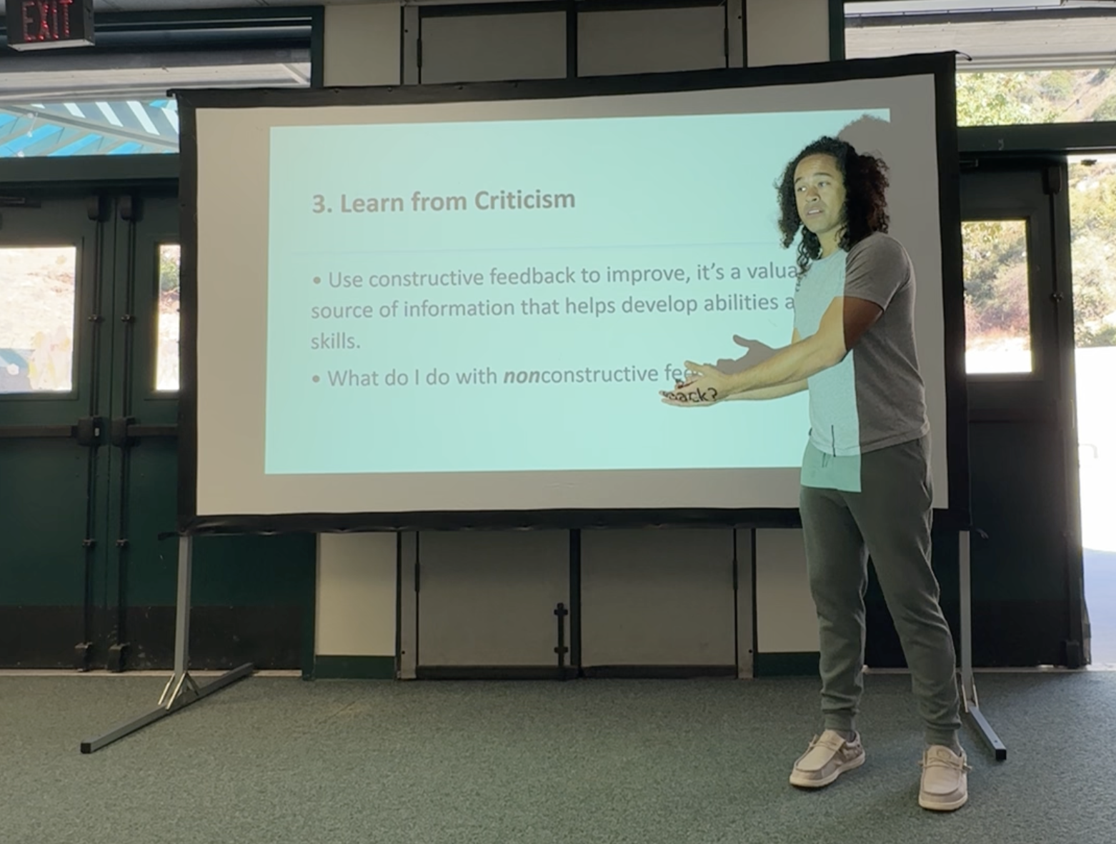 Person giving a presentation about learning from criticism, standing next to a screen displaying slide text, indoors with large windows showing trees outside.
