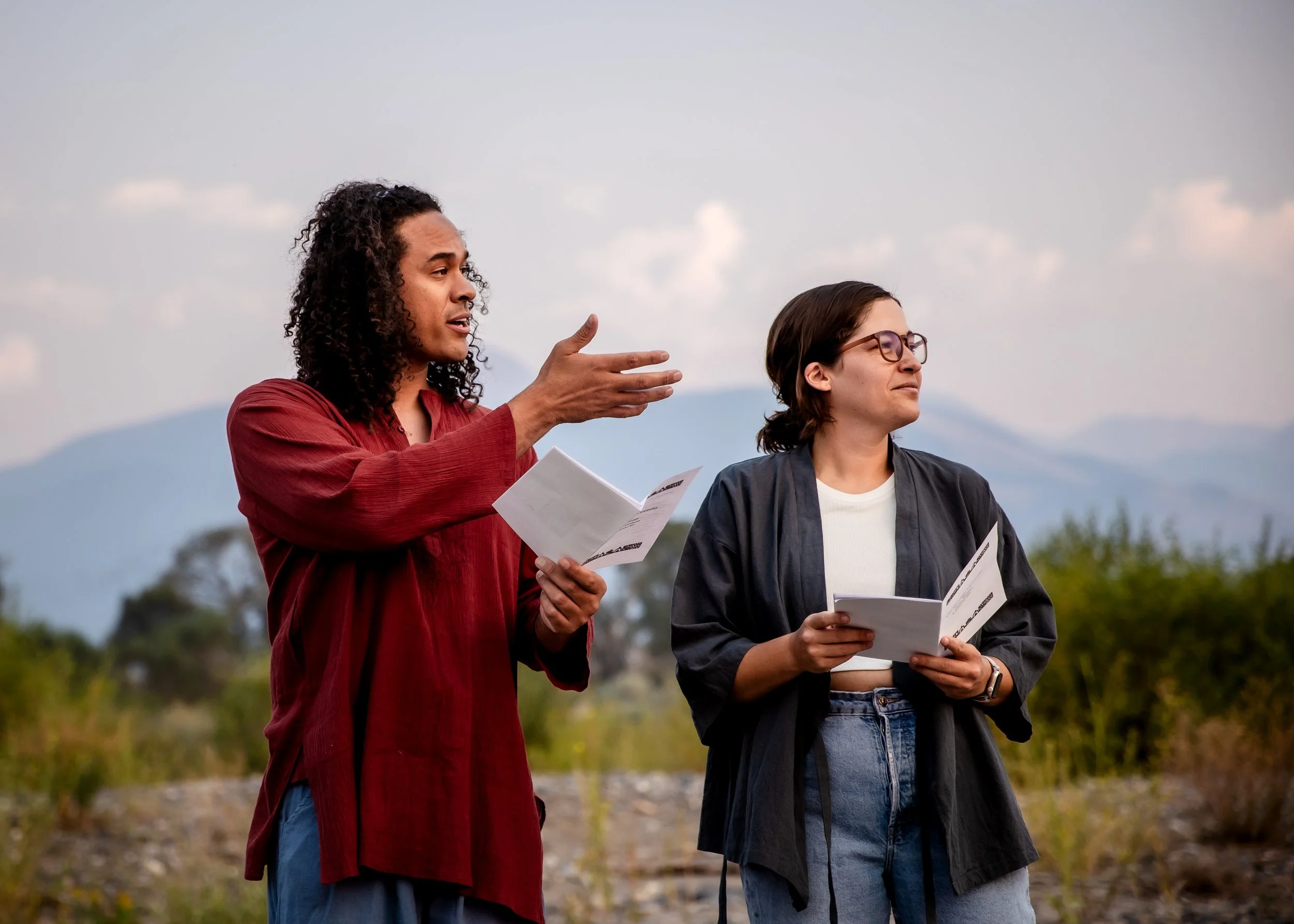 Two people, a man and a woman, standing outdoors in a natural landscape with mountains in the background, engaged in a conversation with the man gesturing with his hand and both holding papers.