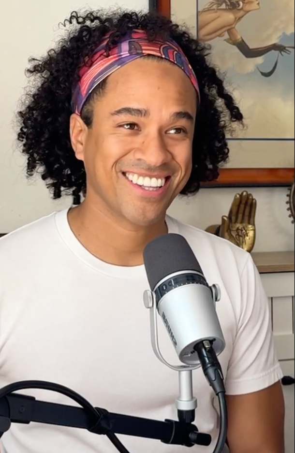 Smiling man with curly hair and a colorful headband, speaking into a microphone during a recording or podcast session.