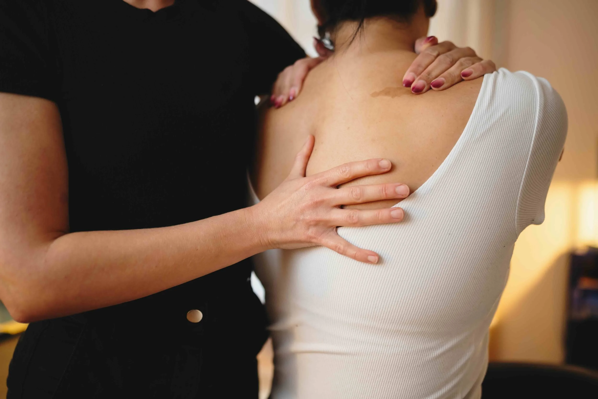 Close-up of two women, one in black shirt and the other in a white sleeveless top, touching their shoulders and back in an embrace.