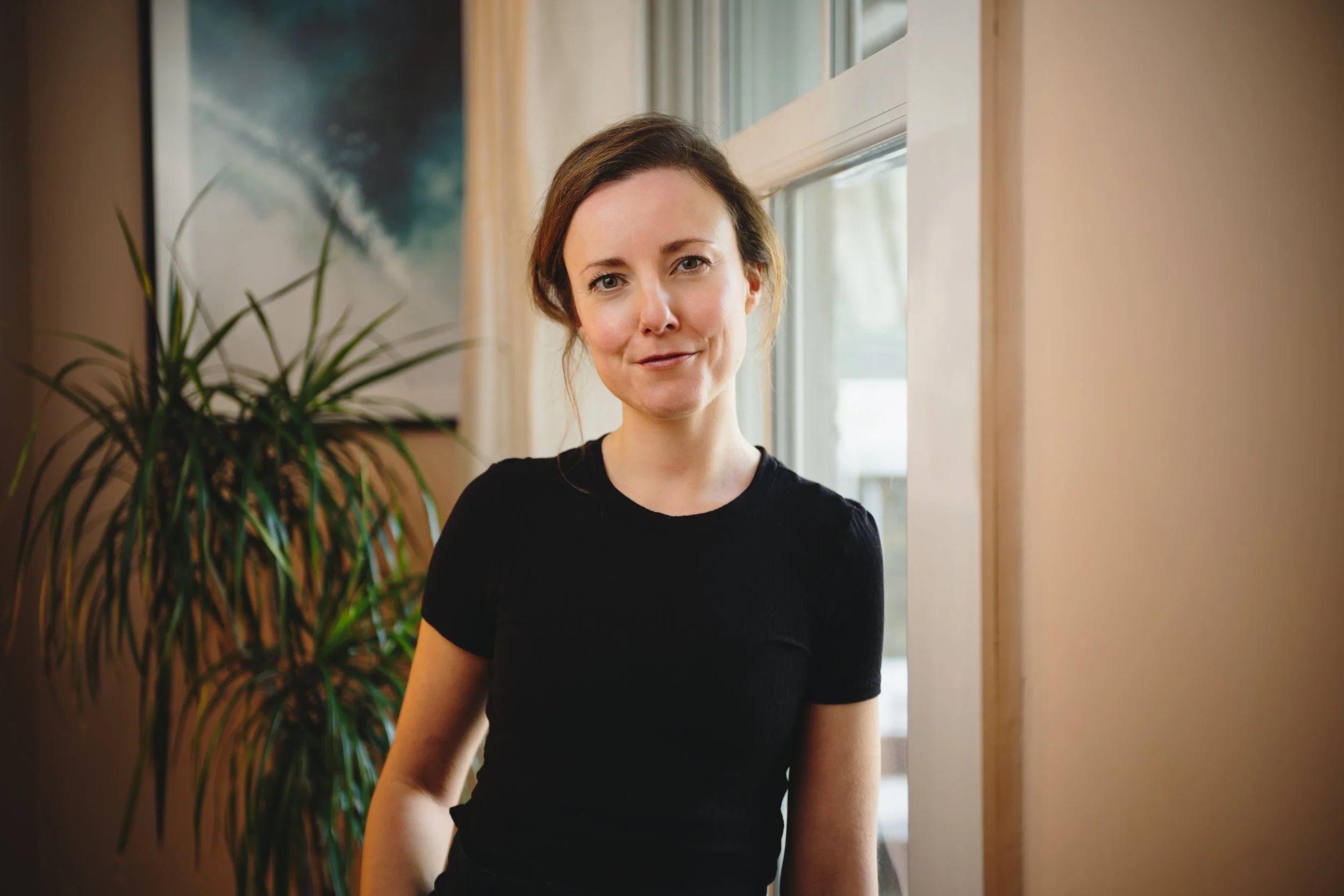 A female Osteopath with shoulder-length brown hair wearing a black shirt, standing indoors near a window with a potted plant and artwork in the background.
