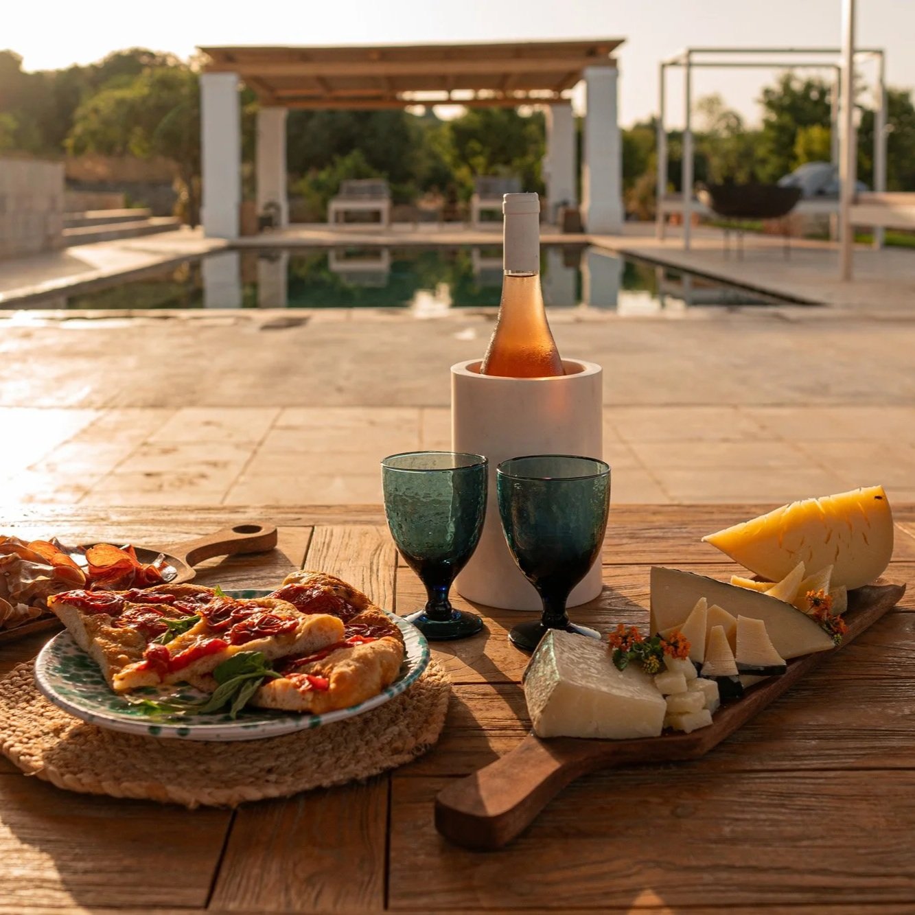 A wooden table set with pizza, assorted cheeses, a bottle of rosé, two wine glasses, and a small plate of snacks, with a swimming pool and outdoor seating area in the background during sunset.