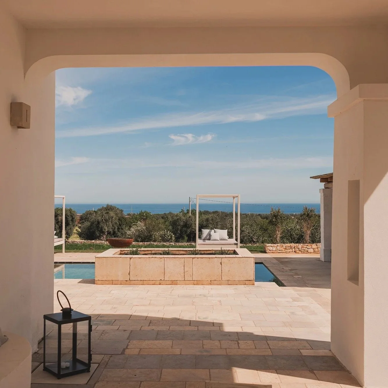 View from a covered patio overlooking a backyard with a swimming pool, two white outdoor beds with pillows, and a view of trees and the ocean in the distance under a blue sky with clouds.