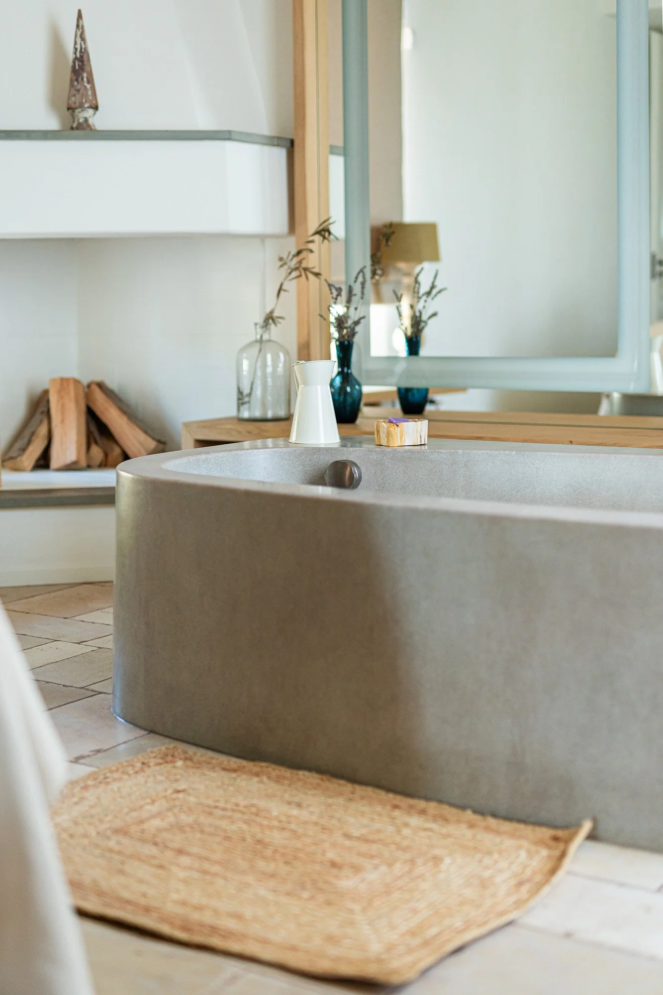 A modern bathroom with a gray bathtub, a beige woven rug, a mirror, and decorative vases with dried plants.
