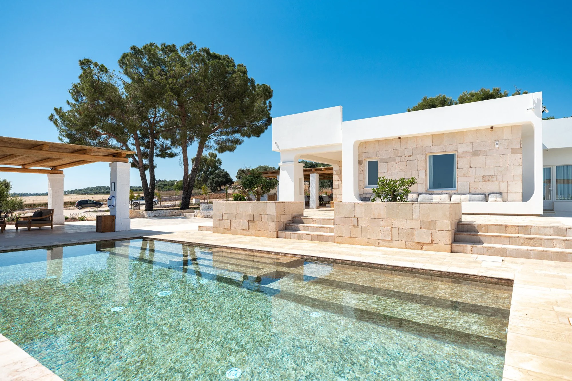 Modern house with stone and white walls, outdoor seating area, and swimming pool under a clear blue sky, with trees and a road in the background.