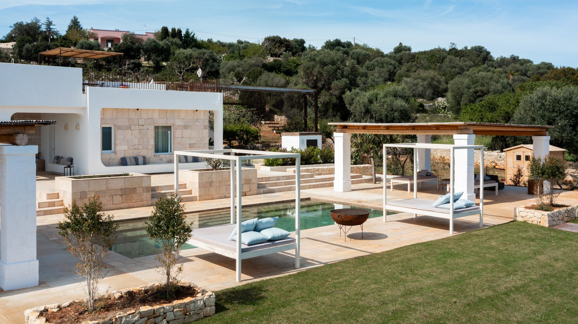 Modern outdoor patio with swimming pool, white lounge beds with blue pillows, and shaded sitting areas surrounded by small trees and greenery in a hillside landscape.
