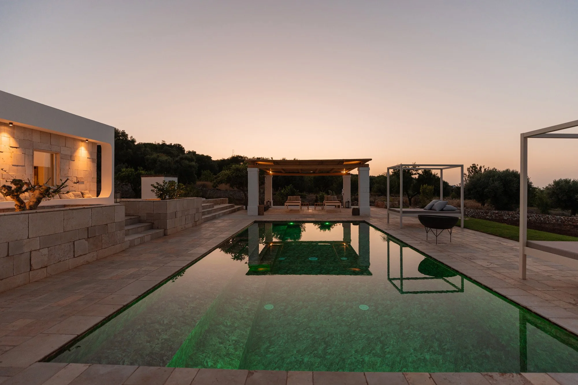 Luxury outdoor pool area at sunset with lounge chairs, shaded cabana, and modern architecture, surrounded by trees and hills.