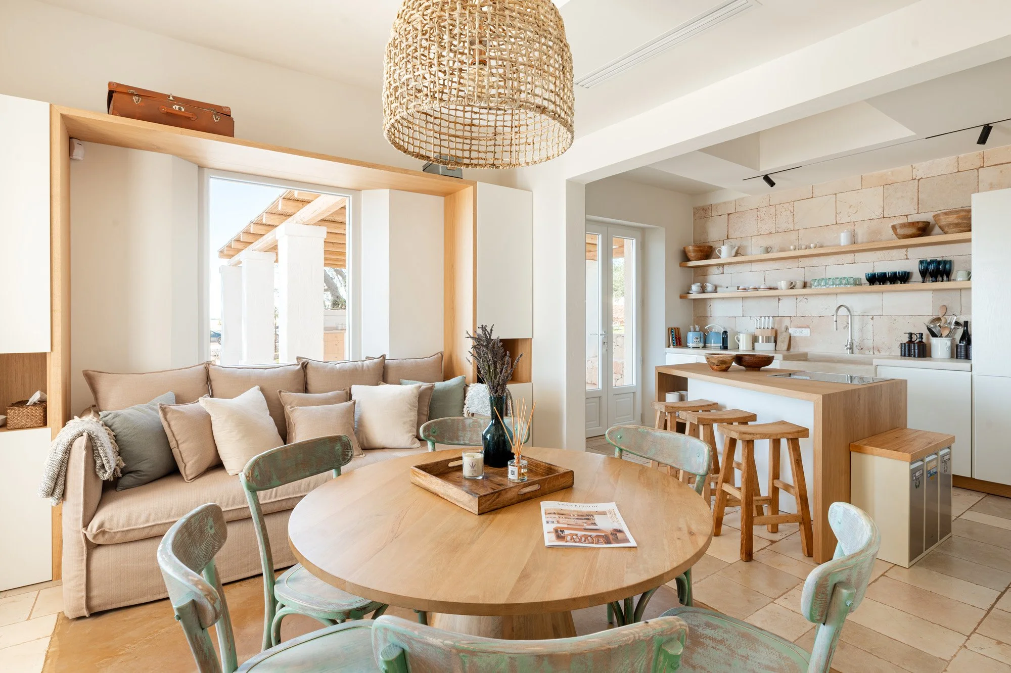 Open-concept living room and kitchen with beige sofa, round dining table with green chairs, wooden bar stools, open shelves with dishes, and natural light from glass doors.