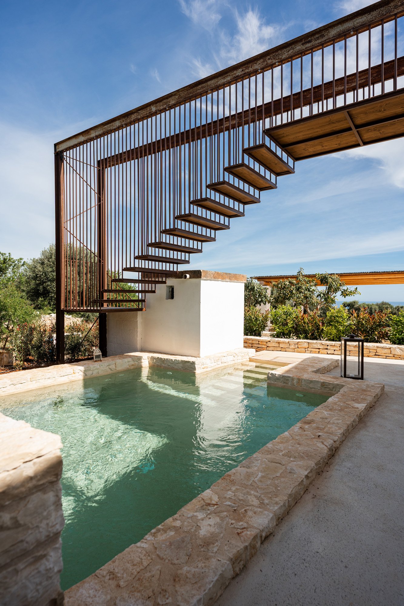 Outdoor swimming pool with stone border and a modern metal staircase structure above, surrounded by greenery and a blue sky with clouds.