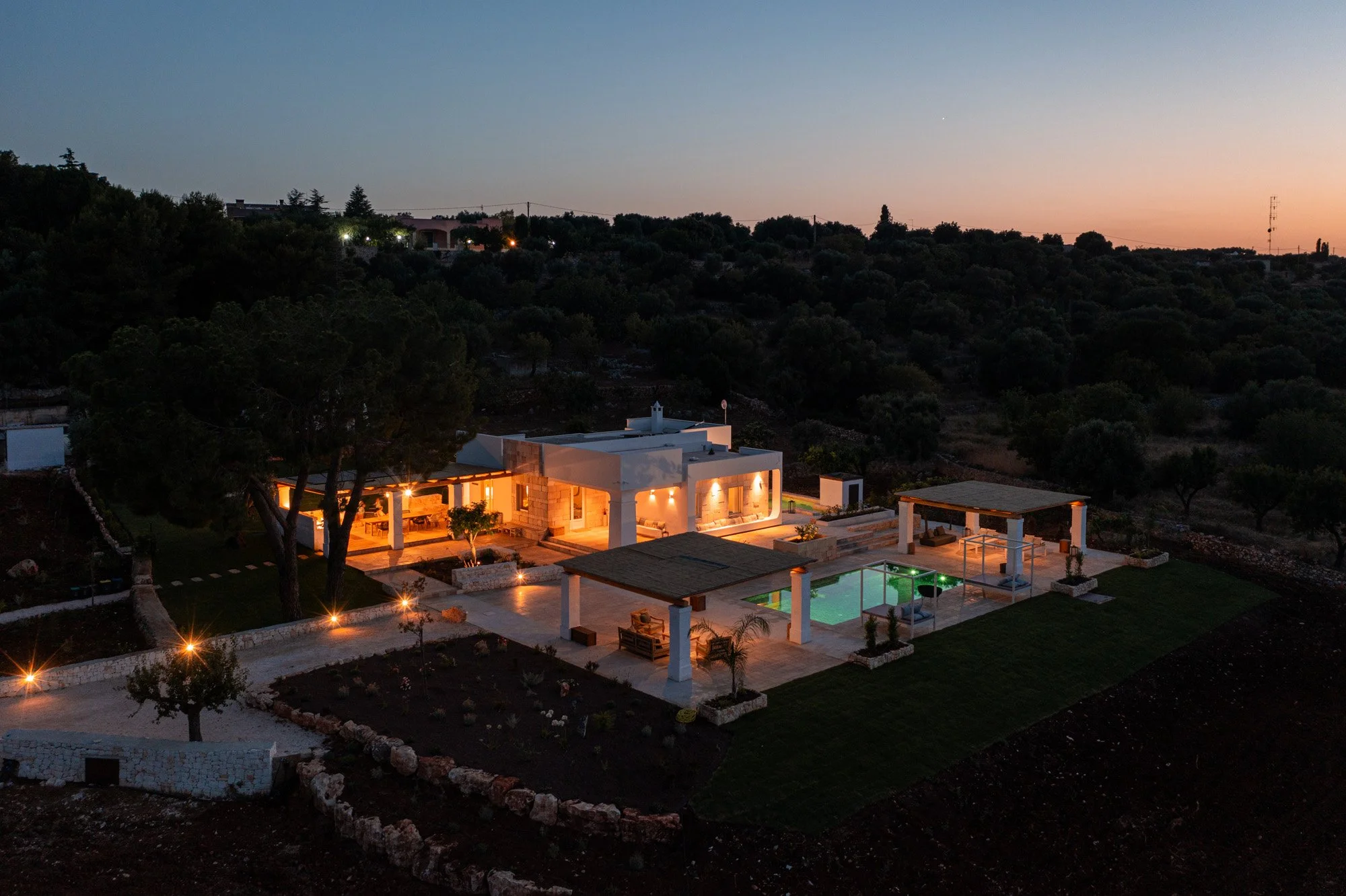 A modern house with an illuminated outdoor area and pool, surrounded by trees and a landscape at dusk.