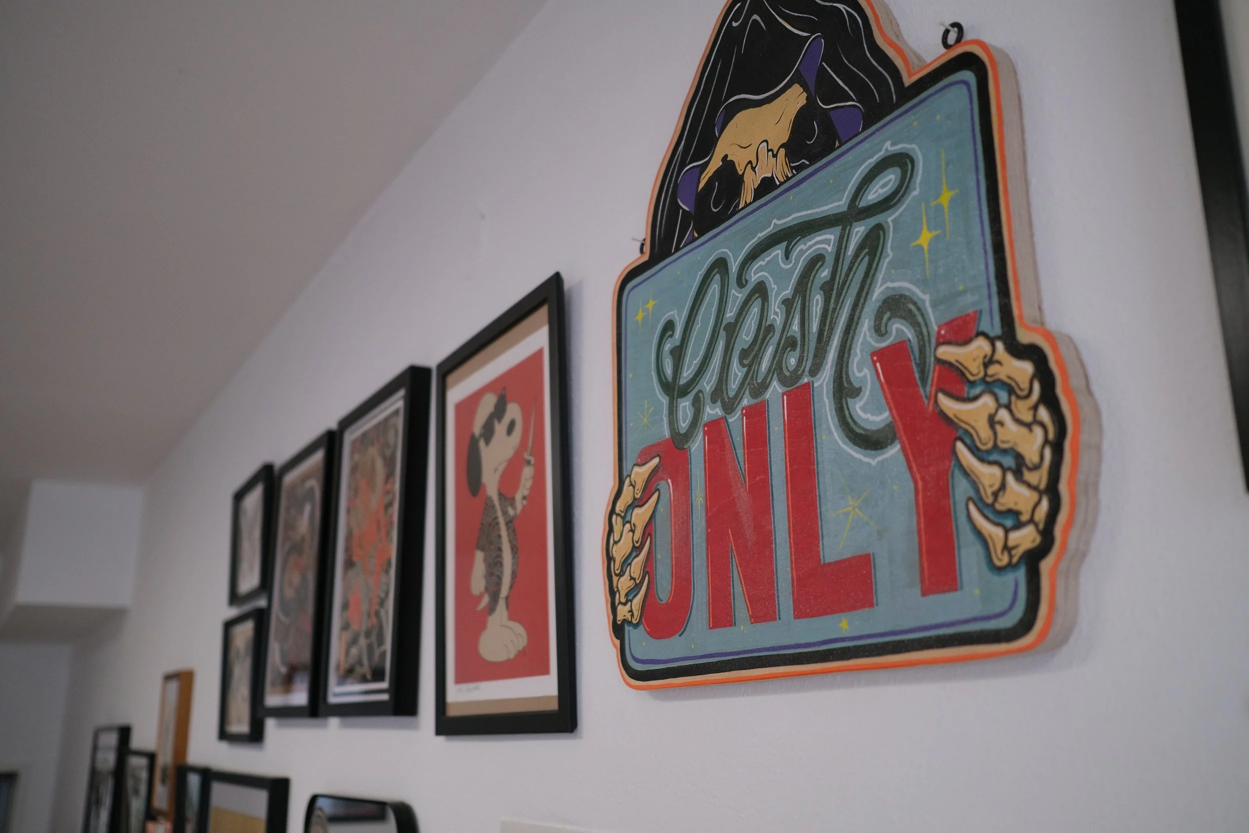 Wand mit mehreren Bilderrahmen, darunter ein Pint-Poster mit dem Schriftzug 'CINNY' und einer Hand, die das Wort hält, sowie ein Bild von einem Tier, vermutlich ein Hund, und eine andere Illustration.