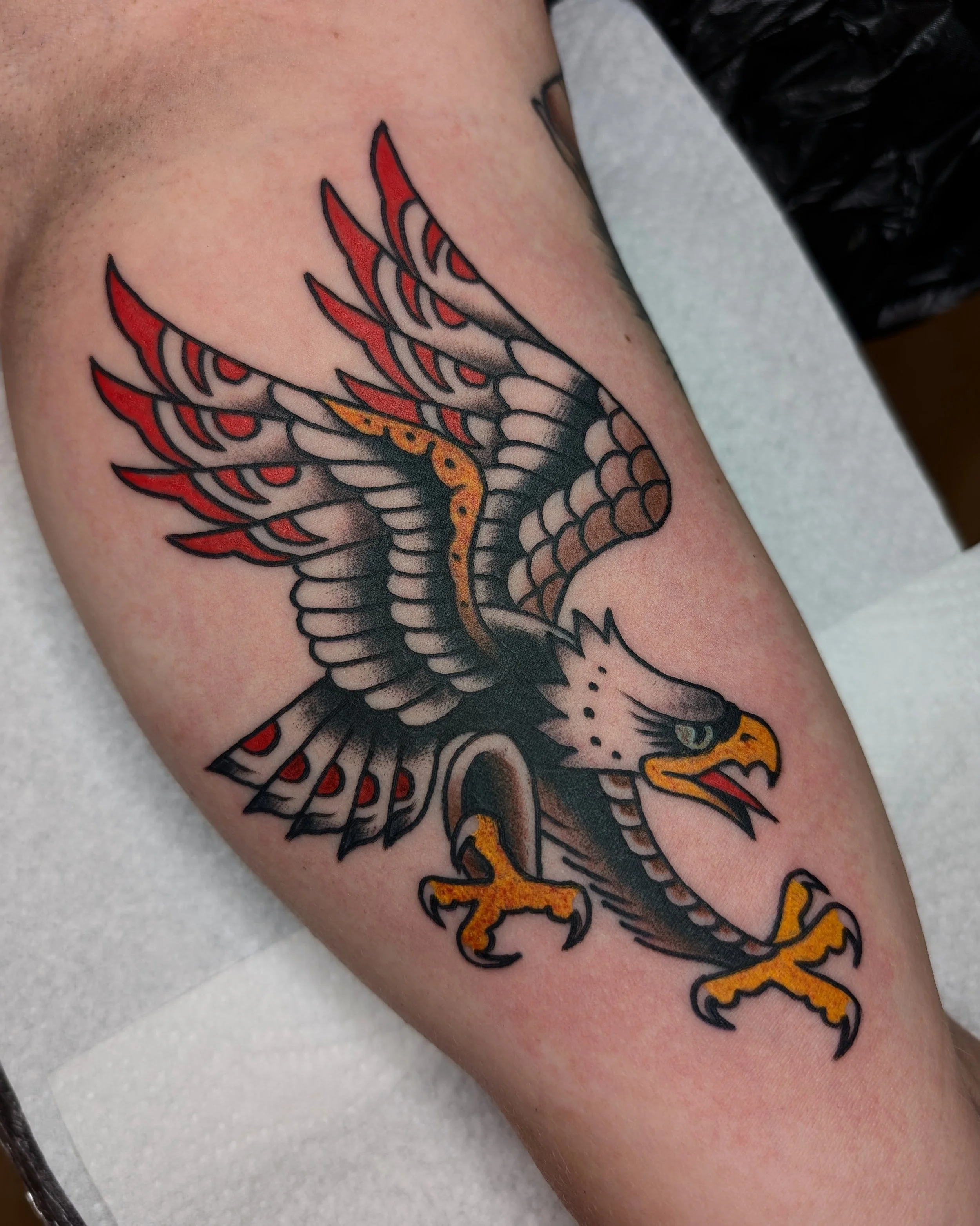 Tattoo eines fliegenden Adlers mit ausgebreiteten Flügeln, gelbem Schnabel und Krallen, schwarze, graue, rote und gelbe Details.