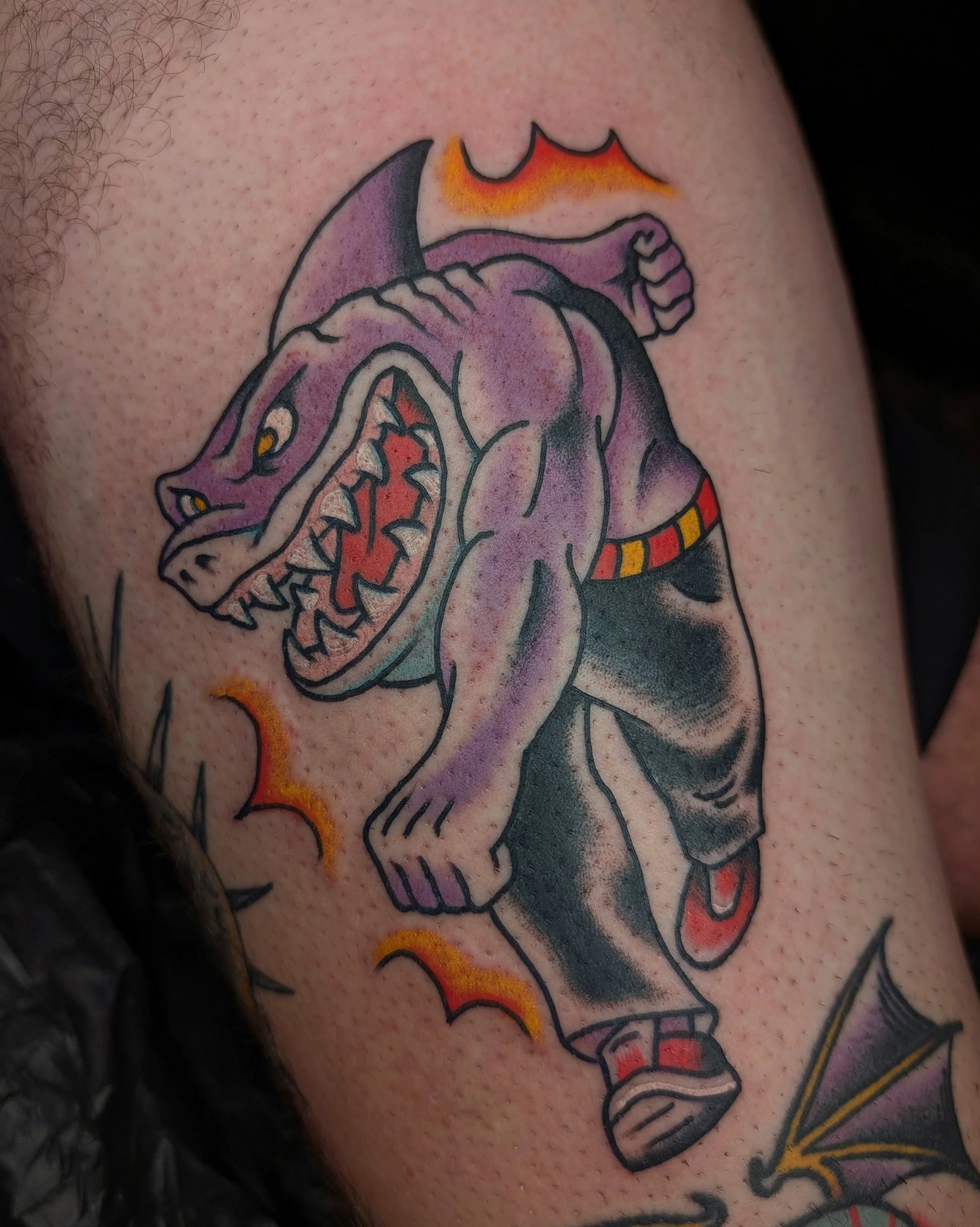 Tattoo eines kämpfenden Monsters, das wie ein Dämon aussieht, mit einem großen Mund voller Zähne, kleinen Flügeln, und in Taschen oder Hosen mit rotem und gelbem Gürtel sowie roten Schuhen. Es ist umgeben von flammenartigen Linien.