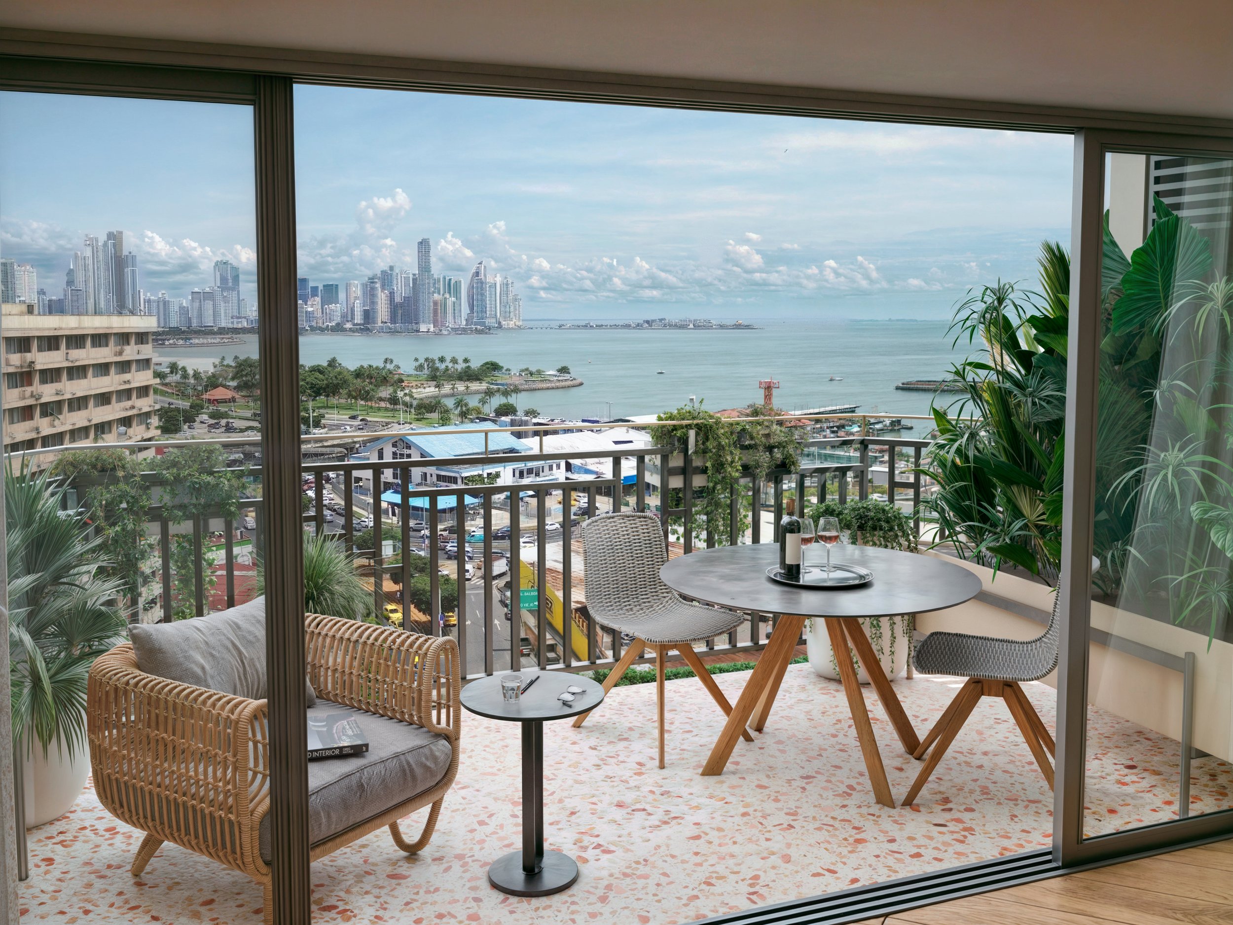 Balcony with a round table, two chairs, sofa, and potted plants, overlooking a city skyline, bay, and boats.