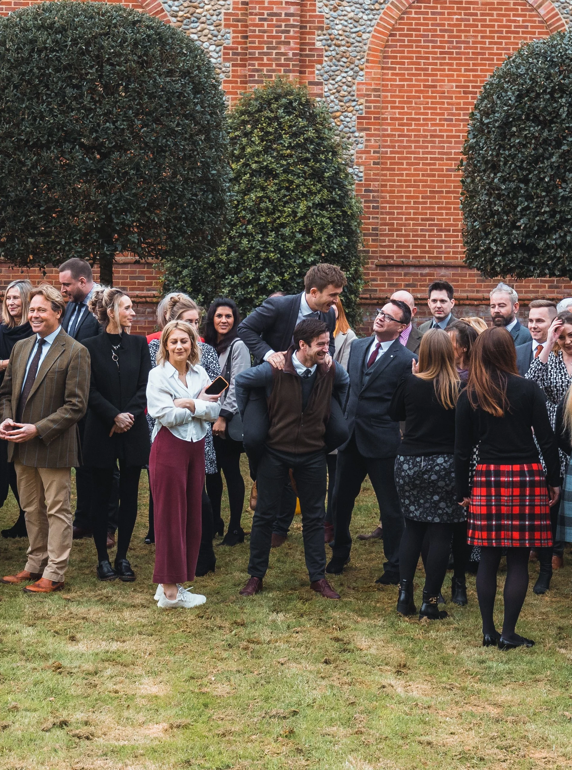 People gathered outdoors in front of a brick wall with arched windows, some are talking while others are smiling, dressed in business or semi-formal attire, with greenery and trees in the background.