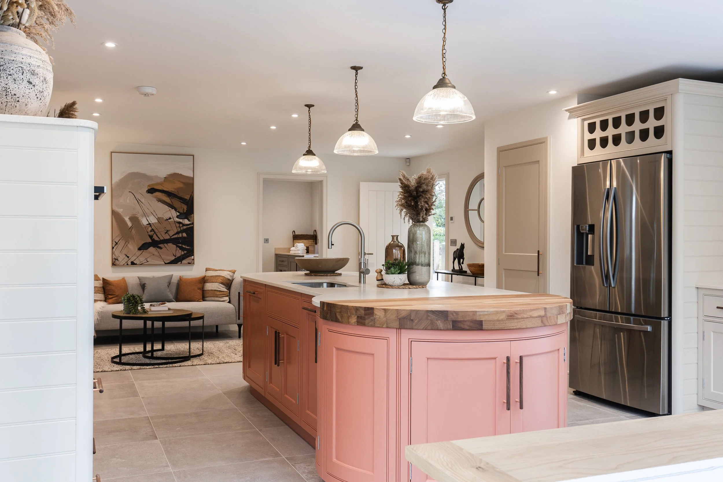 A modern kitchen with pink cabinets, a white countertop, and a stainless steel refrigerator. It features a dark wood tabletop, decorative vases, and lighting fixtures hanging from the ceiling.