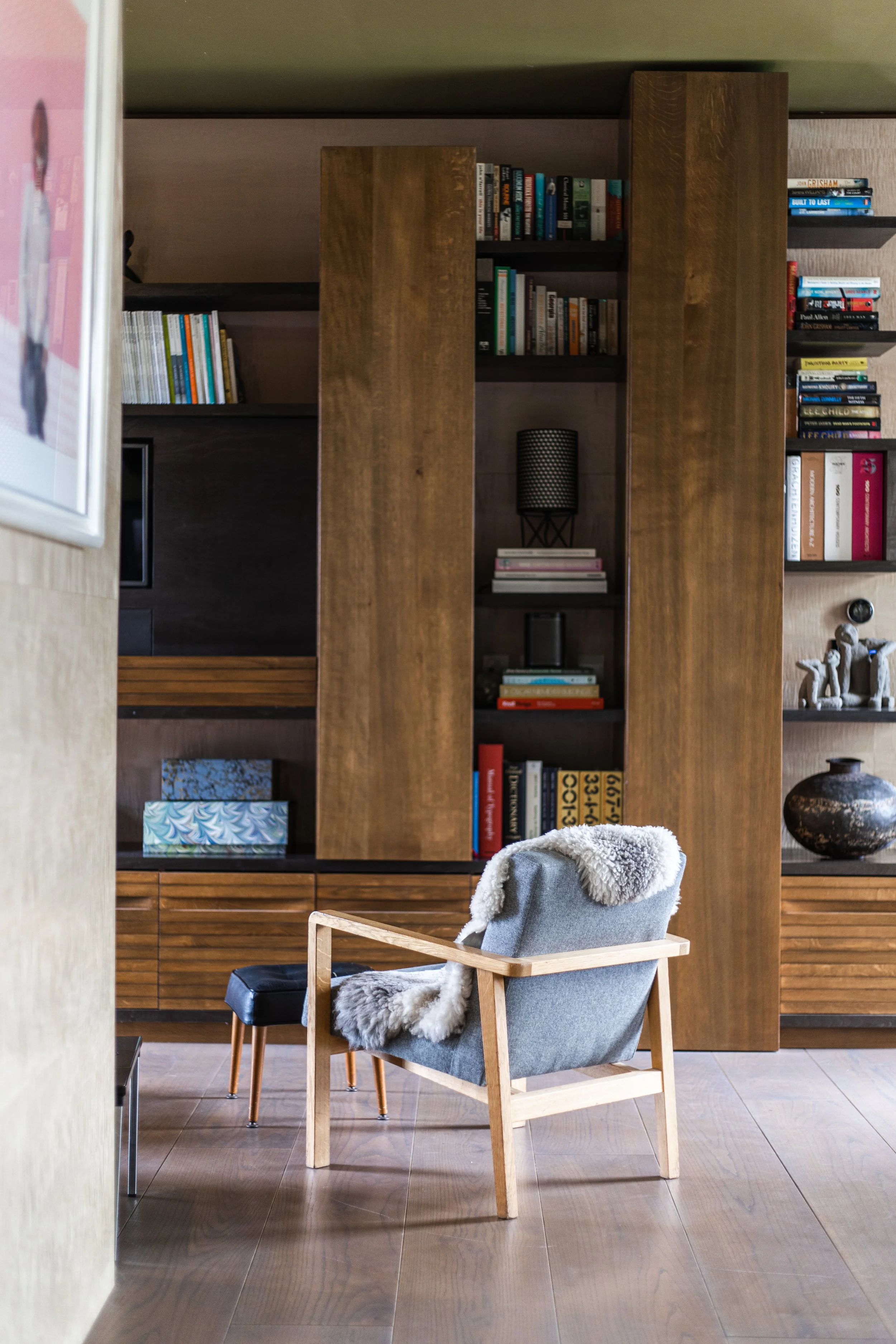 A cozy living room corner featuring a modern wooden armchair with a gray cushion and a plush throw blanket, positioned in front of a tall wooden bookshelf filled with books and decorative items.