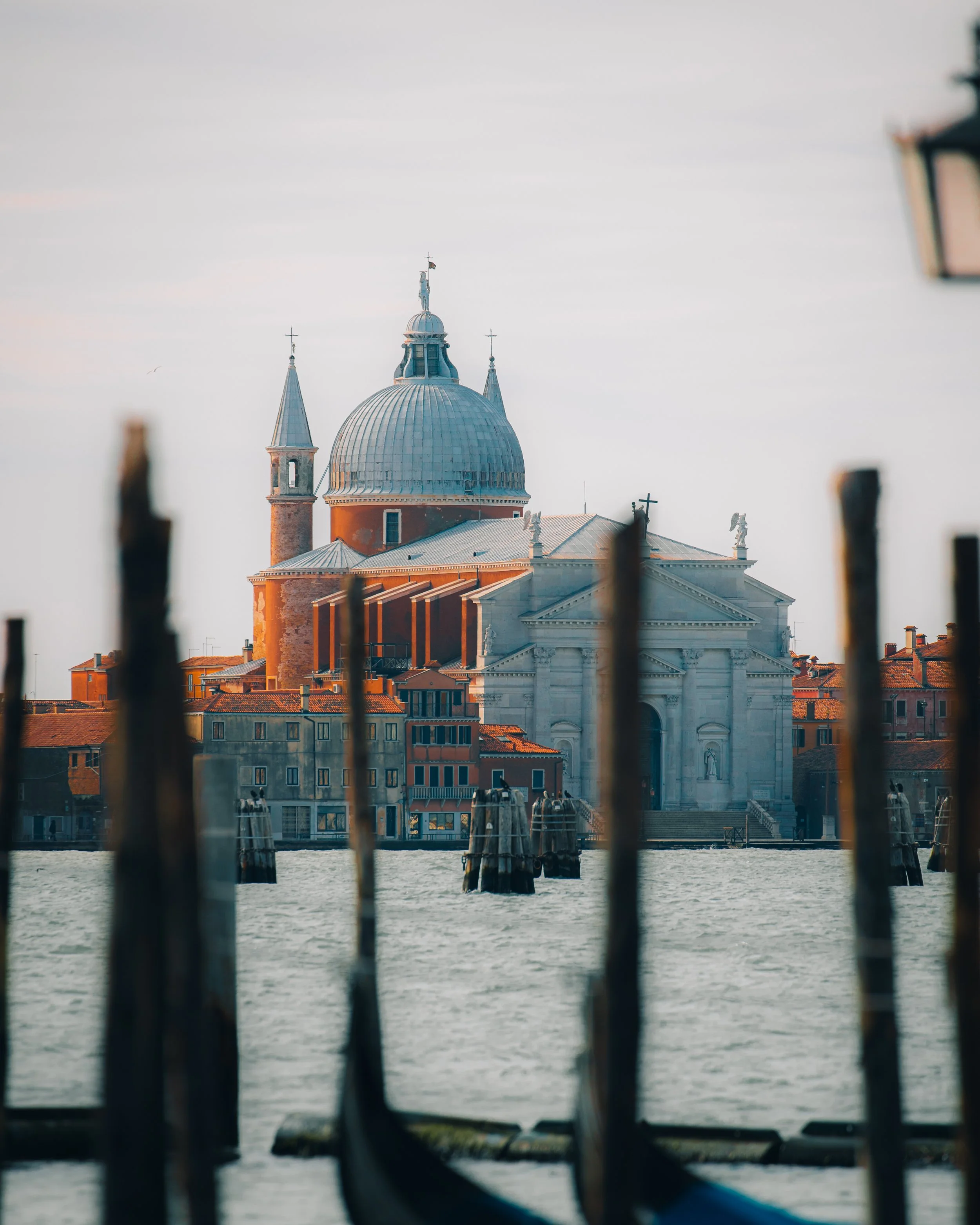 View of a church with a large dome and smaller spires, seen through a fence along a waterway in Venice, Italy.