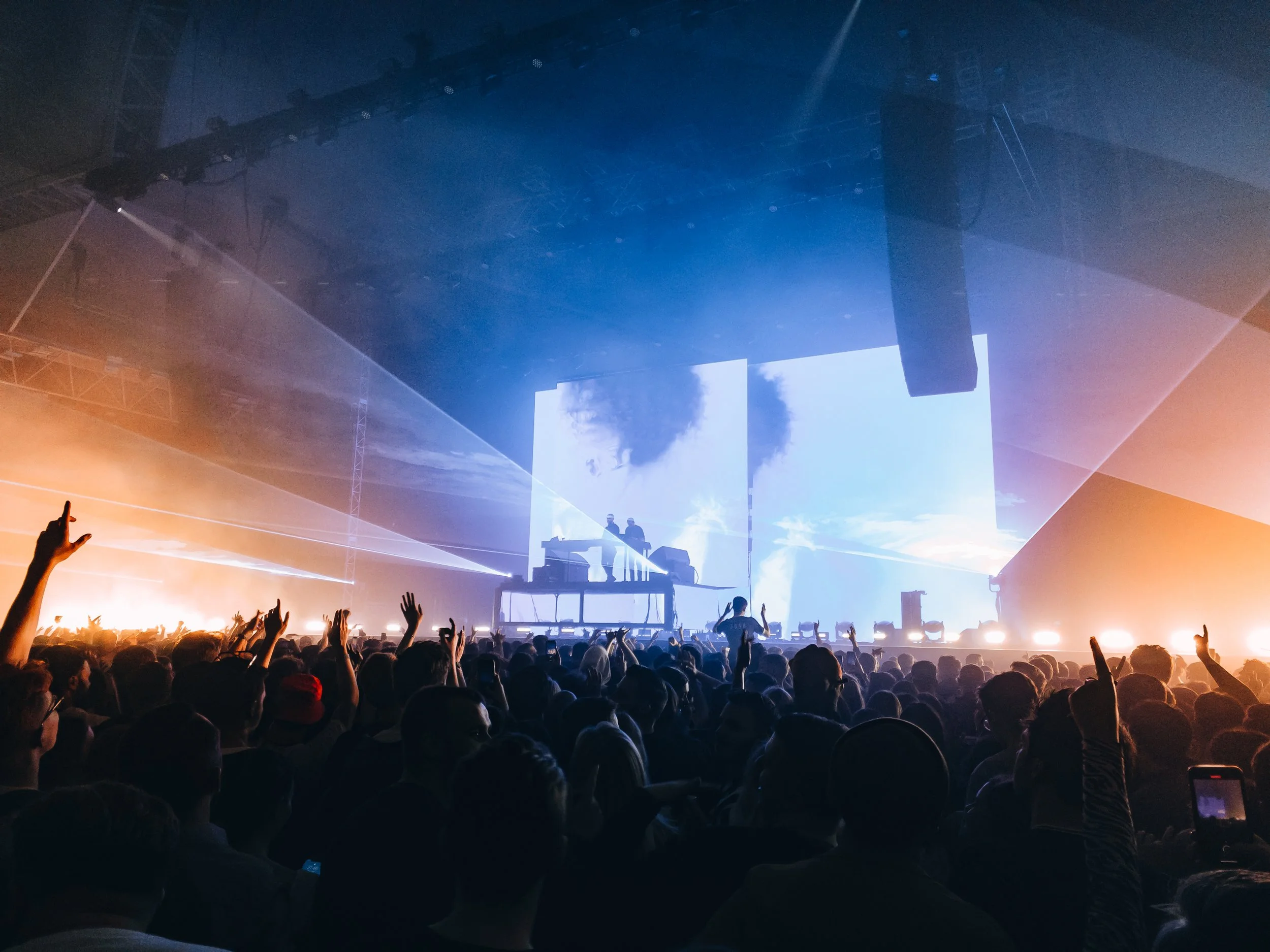 Crowd of concert-goers with hands raised, stage with large screens displaying clouds, DJ on elevated platform, blue and orange lighting.