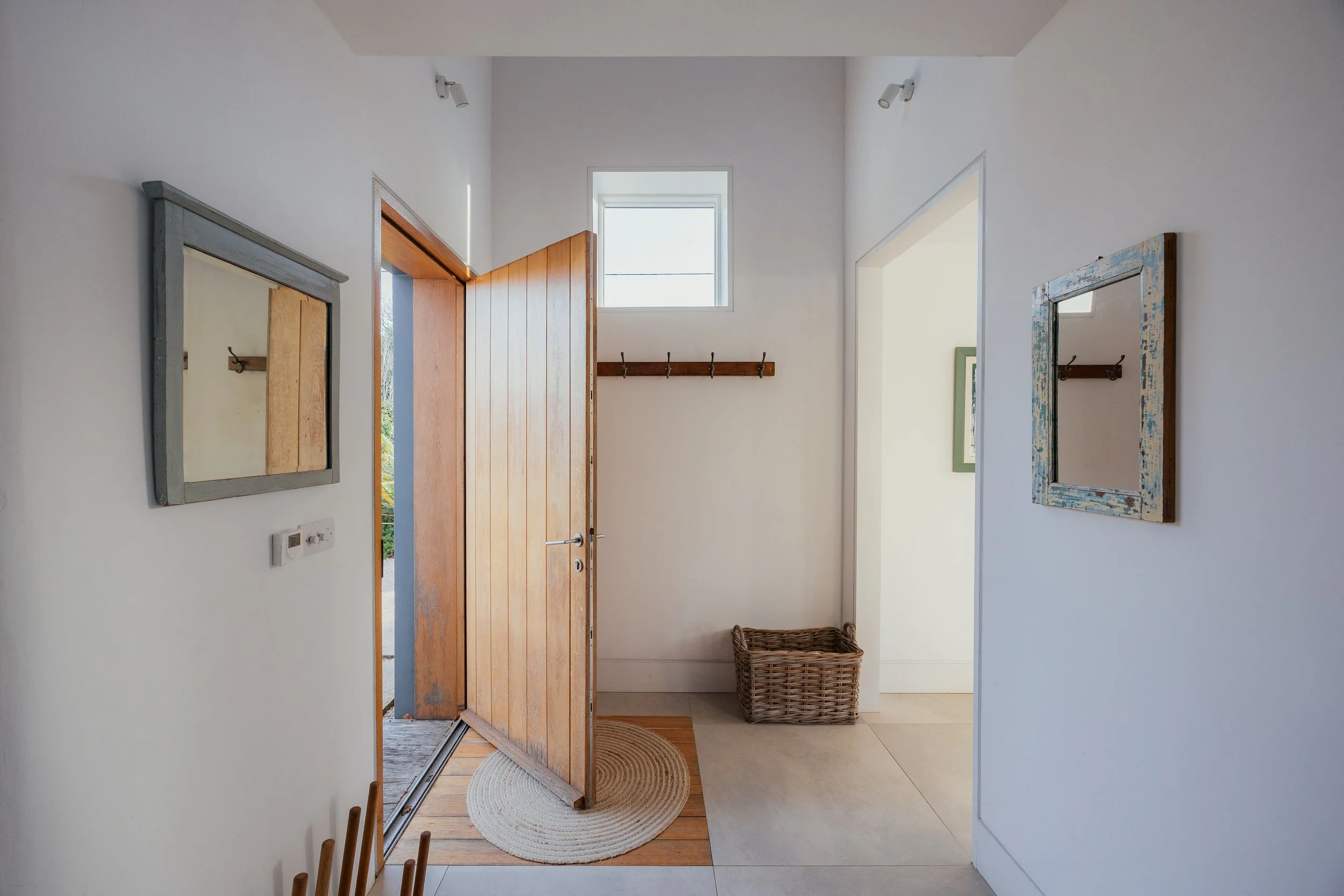 Entrance hall with open wooden door, woven basket, and wall mirrors, bright natural light, white walls.