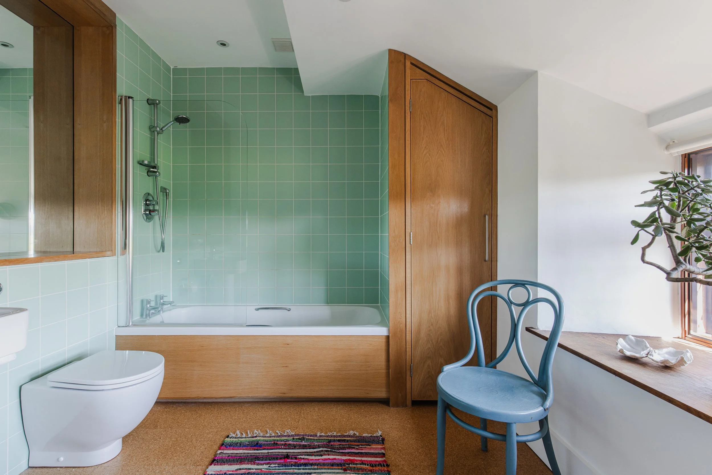 A bathroom with green tiled walls, a bathtub with a wooden panel, a partially visible toilet, a mirror cabinet, a wooden door, a blue chair, a windowsill with decorative shells, and a potted plant