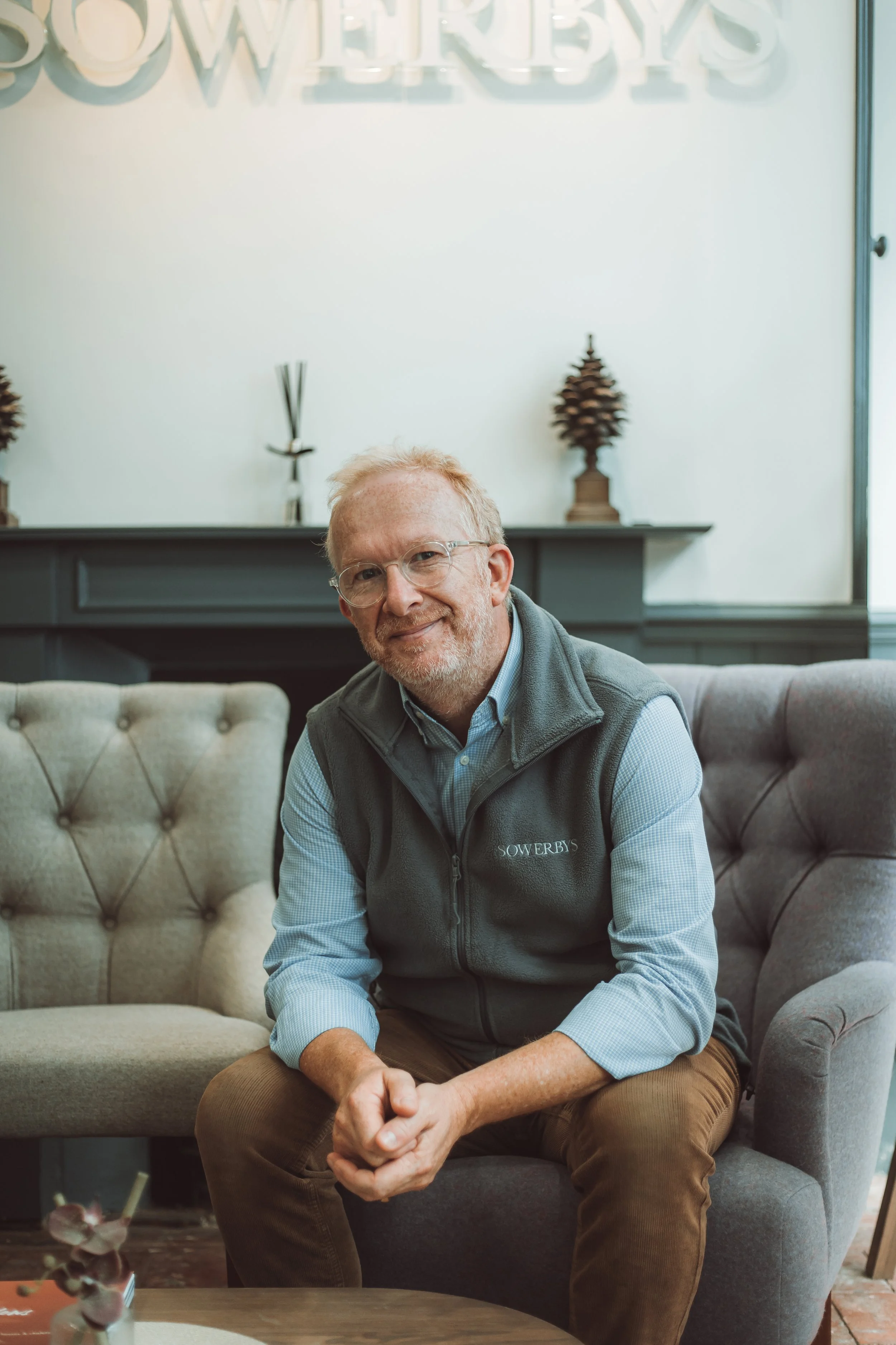 A man with glasses, light-colored hair, and a beard, sitting on a gray tufted armchair indoors. He is smiling, wearing a blue checkered shirt and a dark gray vest with the word 'SOWERBYS' embroidered on it. Behind him is a dark gray fireplace mantel 