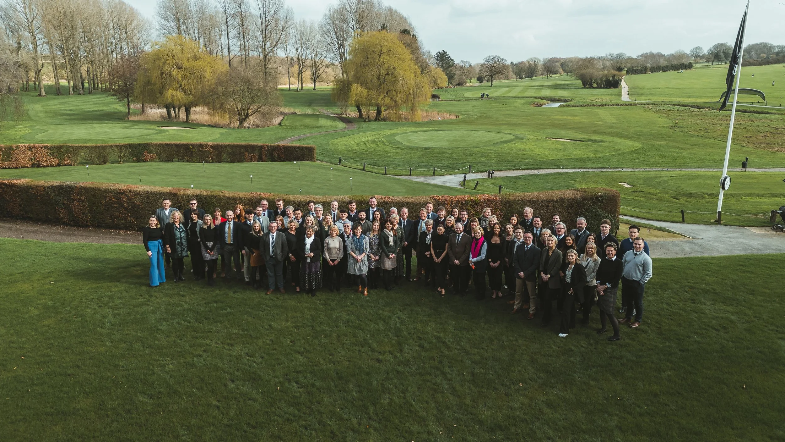 A group of approximately 50 people standing together outdoors on a grassy area at a golf course, with lush green fairways, trees, and a flag on a golf hole in the background.