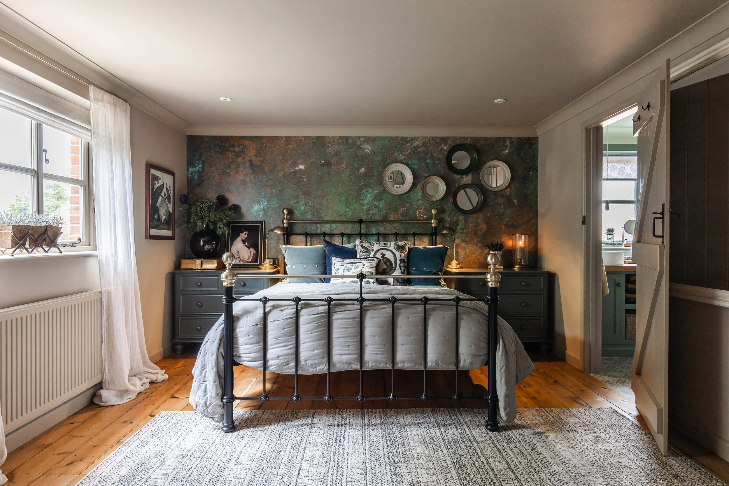 A bedroom with a metallic bed frame, blue and white pillows, dark green nightstands, and a textured accent wall with mirrors.