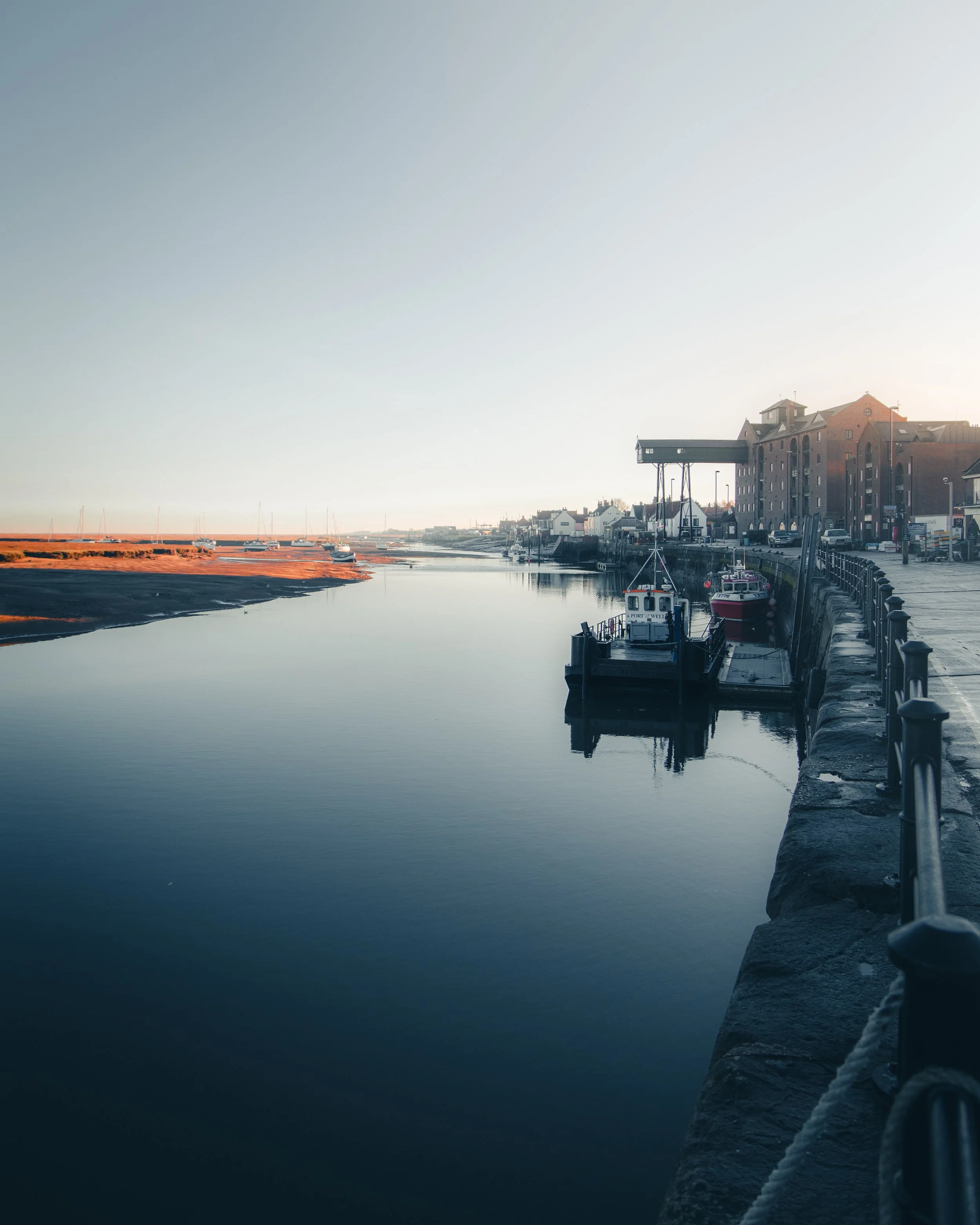 A peaceful harbor scene with boats docked along a calm waterway, buildings lining the street on the right, and a clear sky overhead.