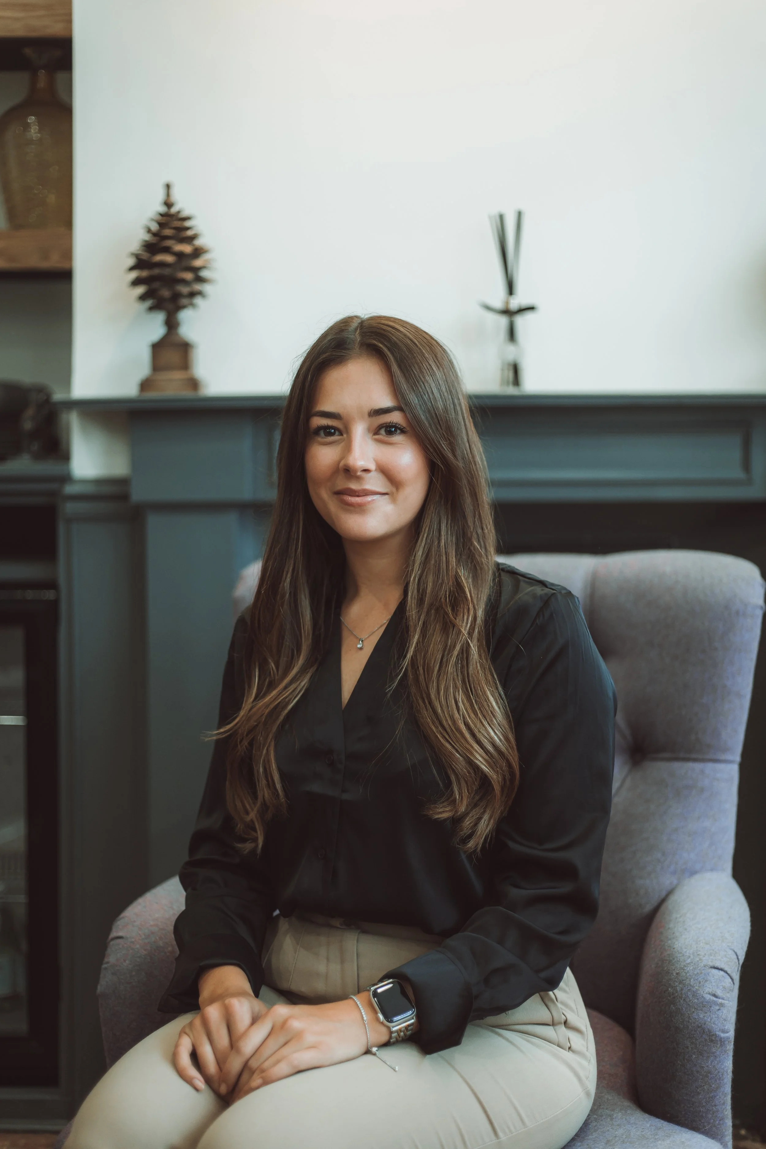 A young woman with long brown hair sitting on a light gray armchair in a room with a dark gray fireplace mantle behind her. She is smiling slightly, wearing a black silk blouse, beige pants, a silver watch, and a delicate necklace.