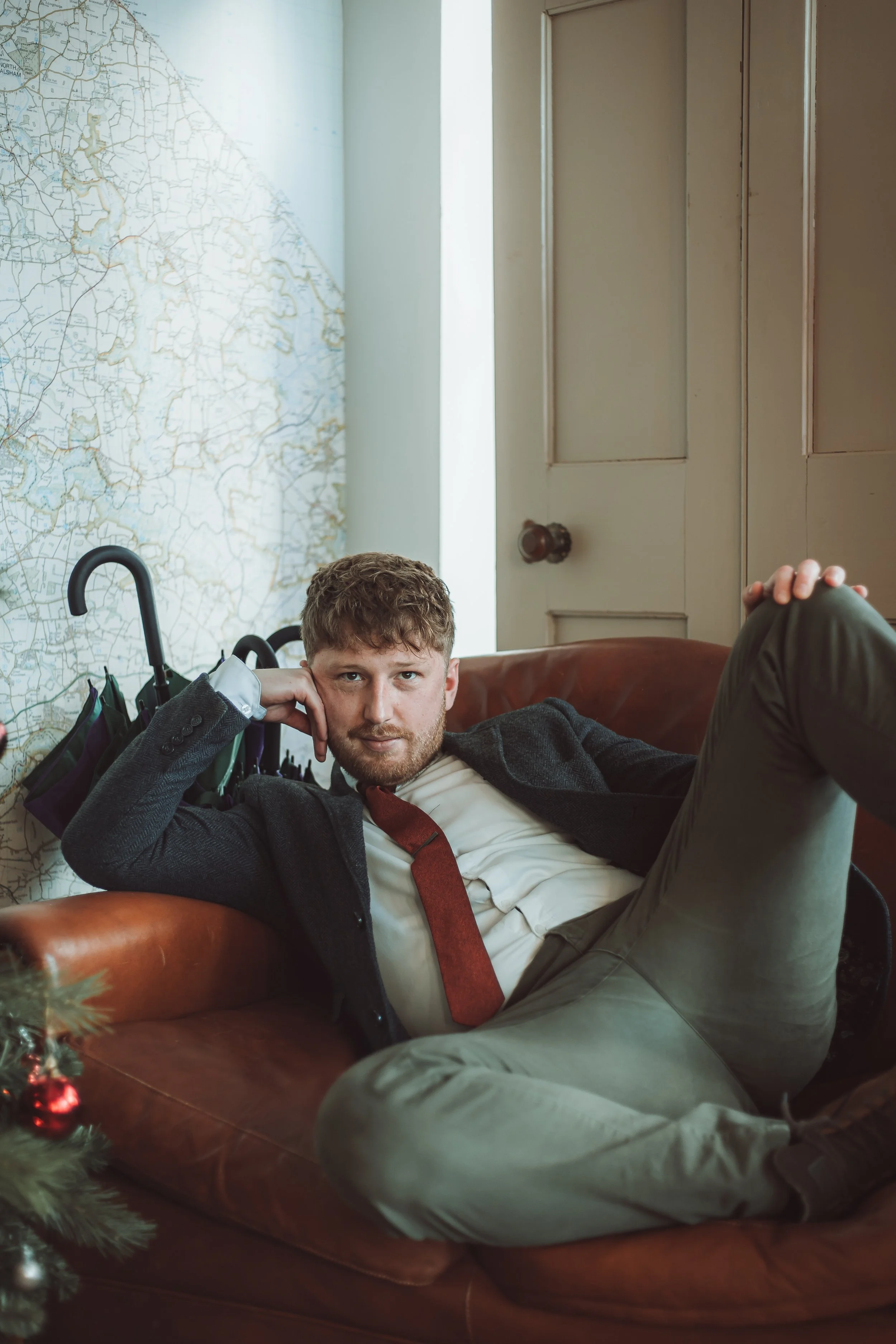 A man with a beard and curly hair lying on a brown leather couch, wearing a gray blazer, white shirt, red tie, and light-colored pants, with his head resting on his hand, in a room with a large map on the wall and a closed door in the background.