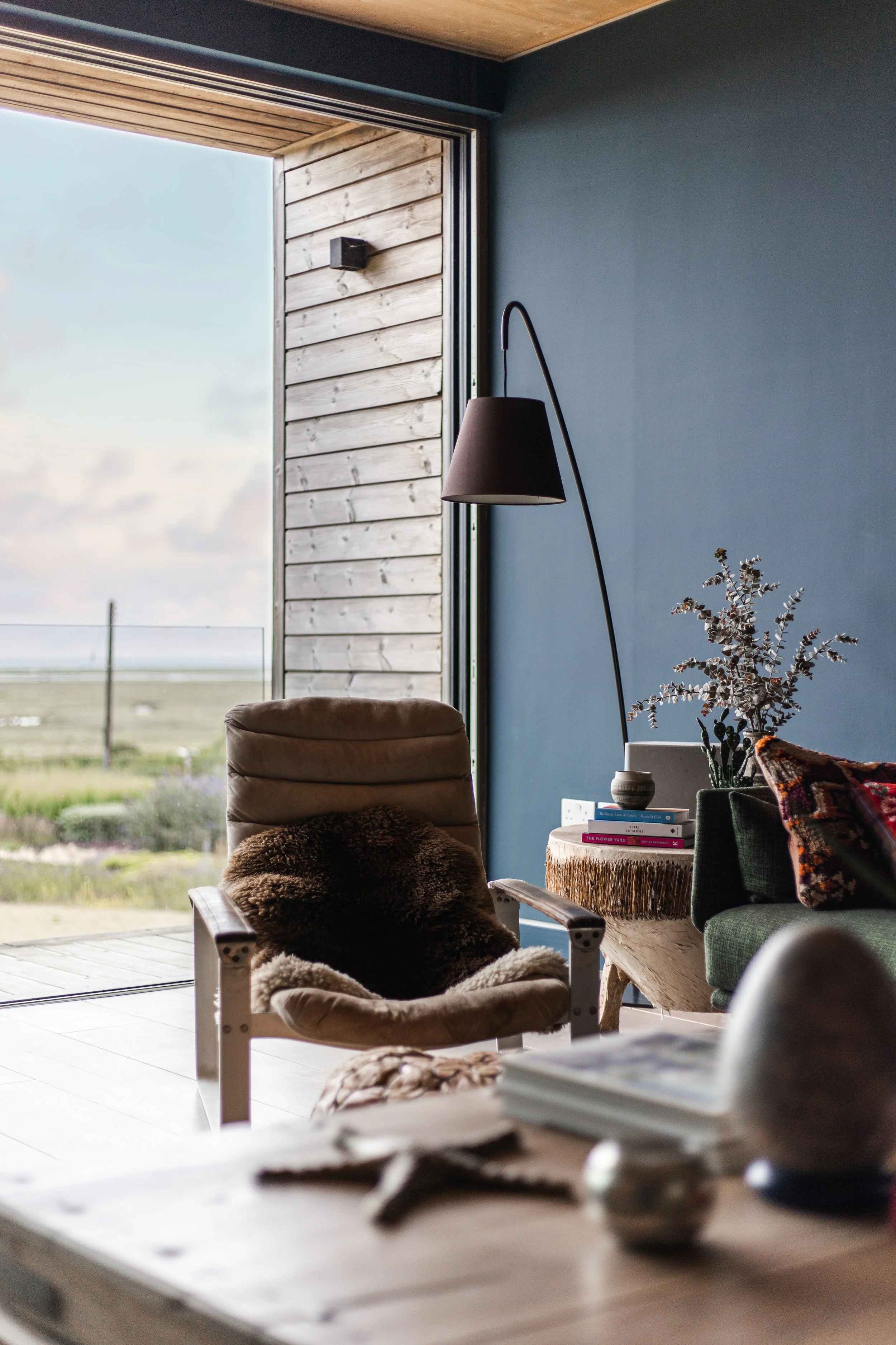 Living room with a large window showing a scenic outdoor view, a beige armchair with a brown cushion, a wooden side table with books and a plant, a black floor lamp, and a green armchair with a colorful pillow.