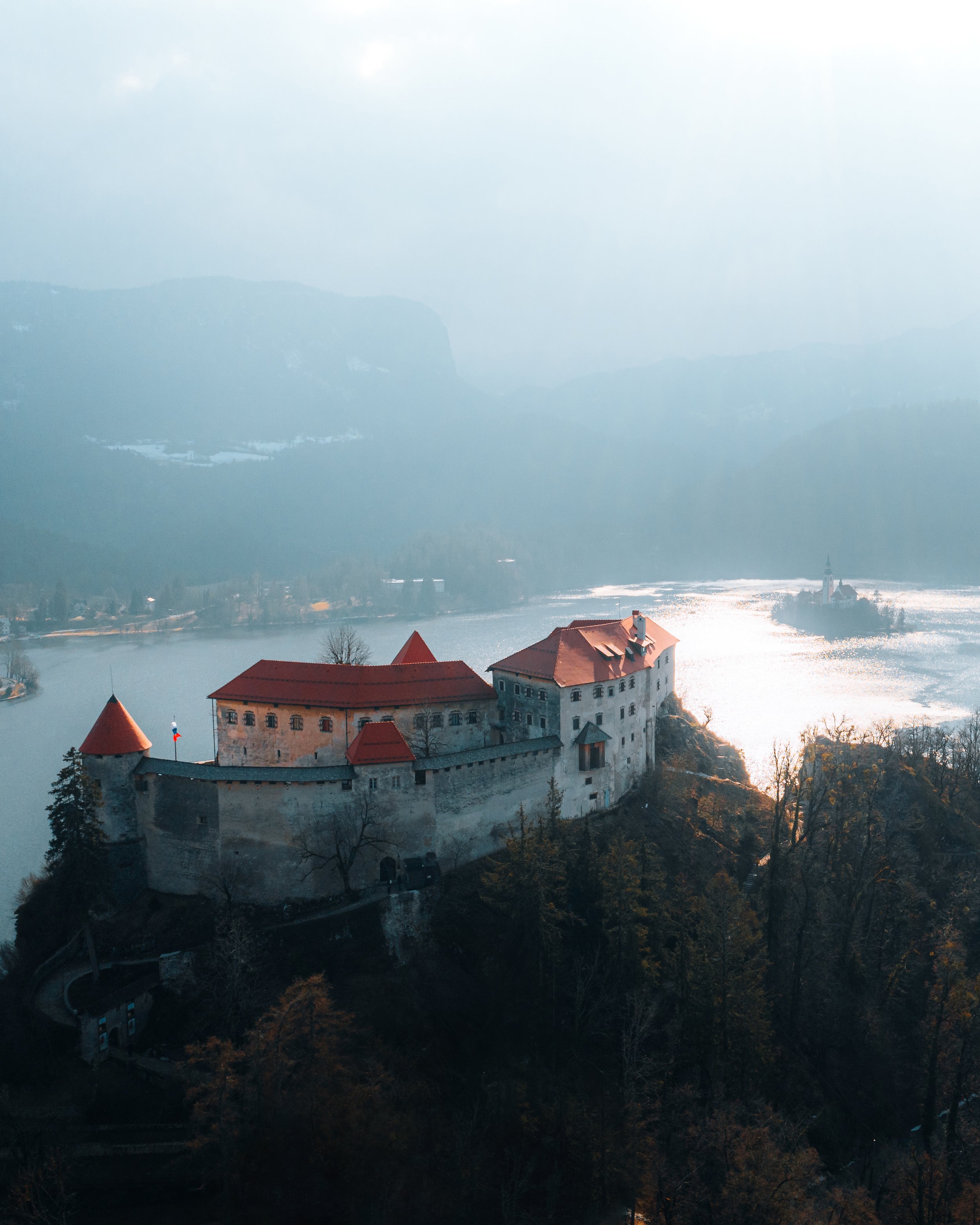 A castle on a hill overlooking a lake, surrounded by trees and mountains in the background.