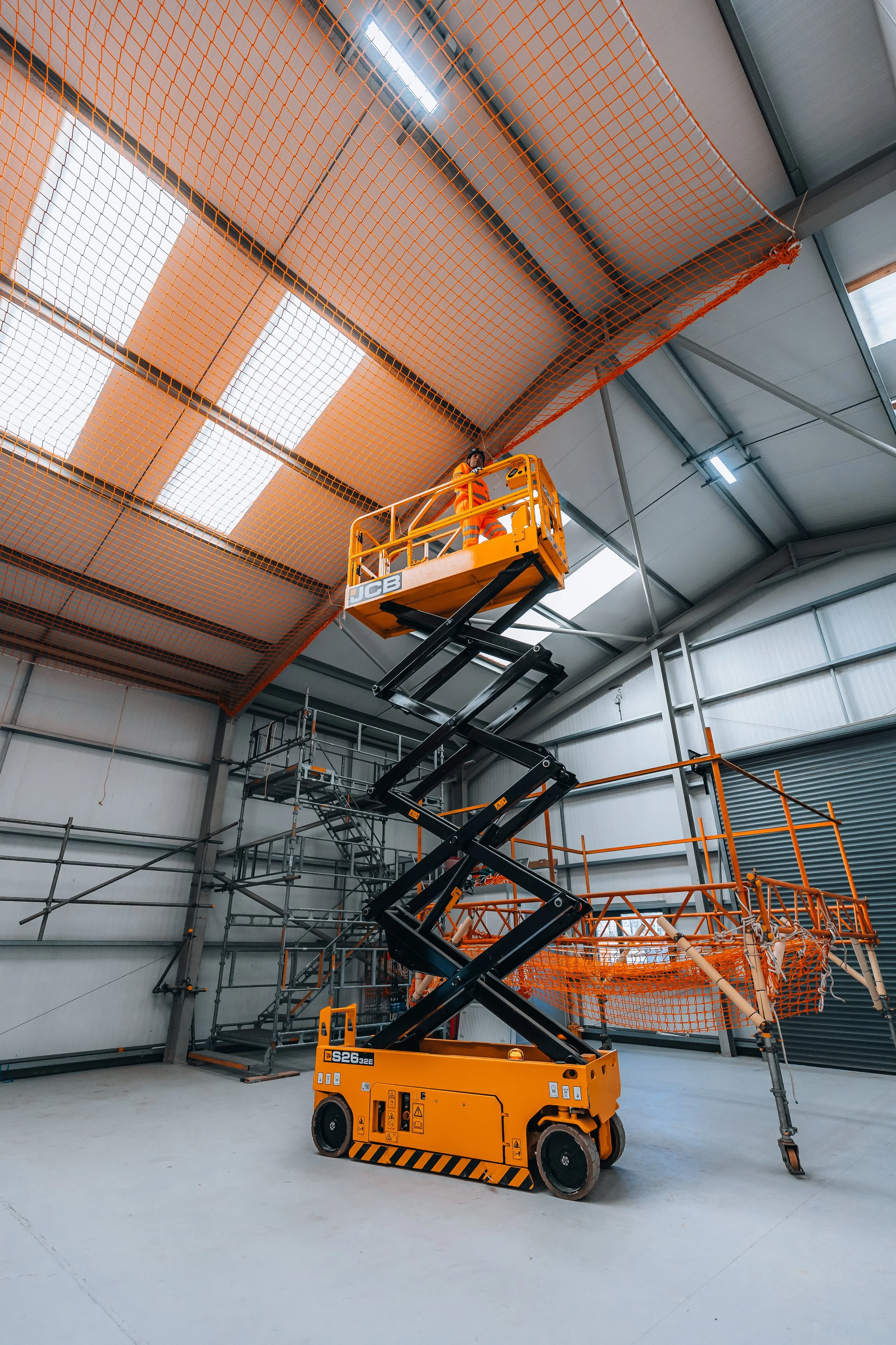 Worker on an orange scissor lift working on the ceiling of a large industrial warehouse, with orange safety netting installed on the ceiling and scaffolding set up along the walls.