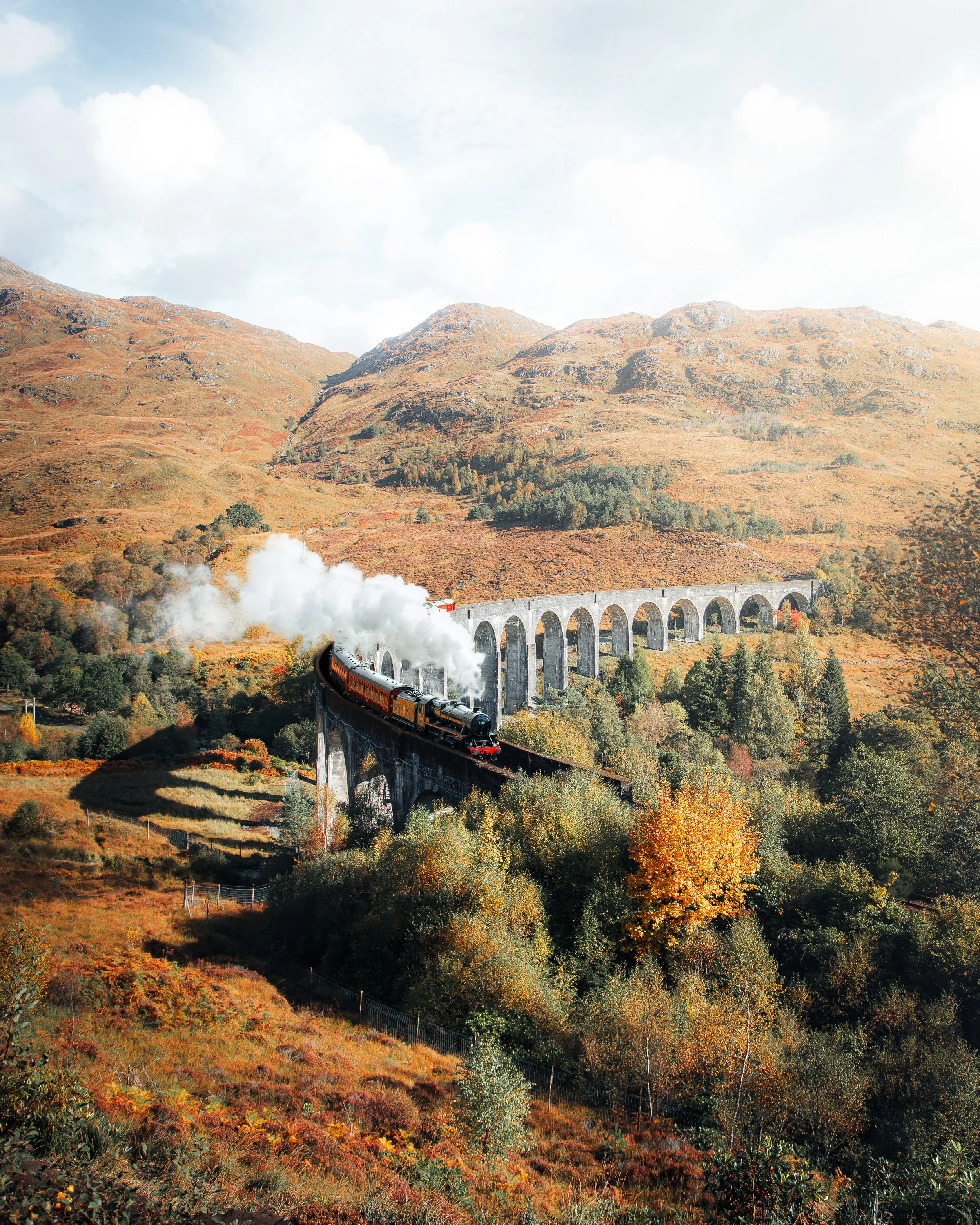 A steam train travels over a curved bridge through a mountainous landscape with autumn foliage.