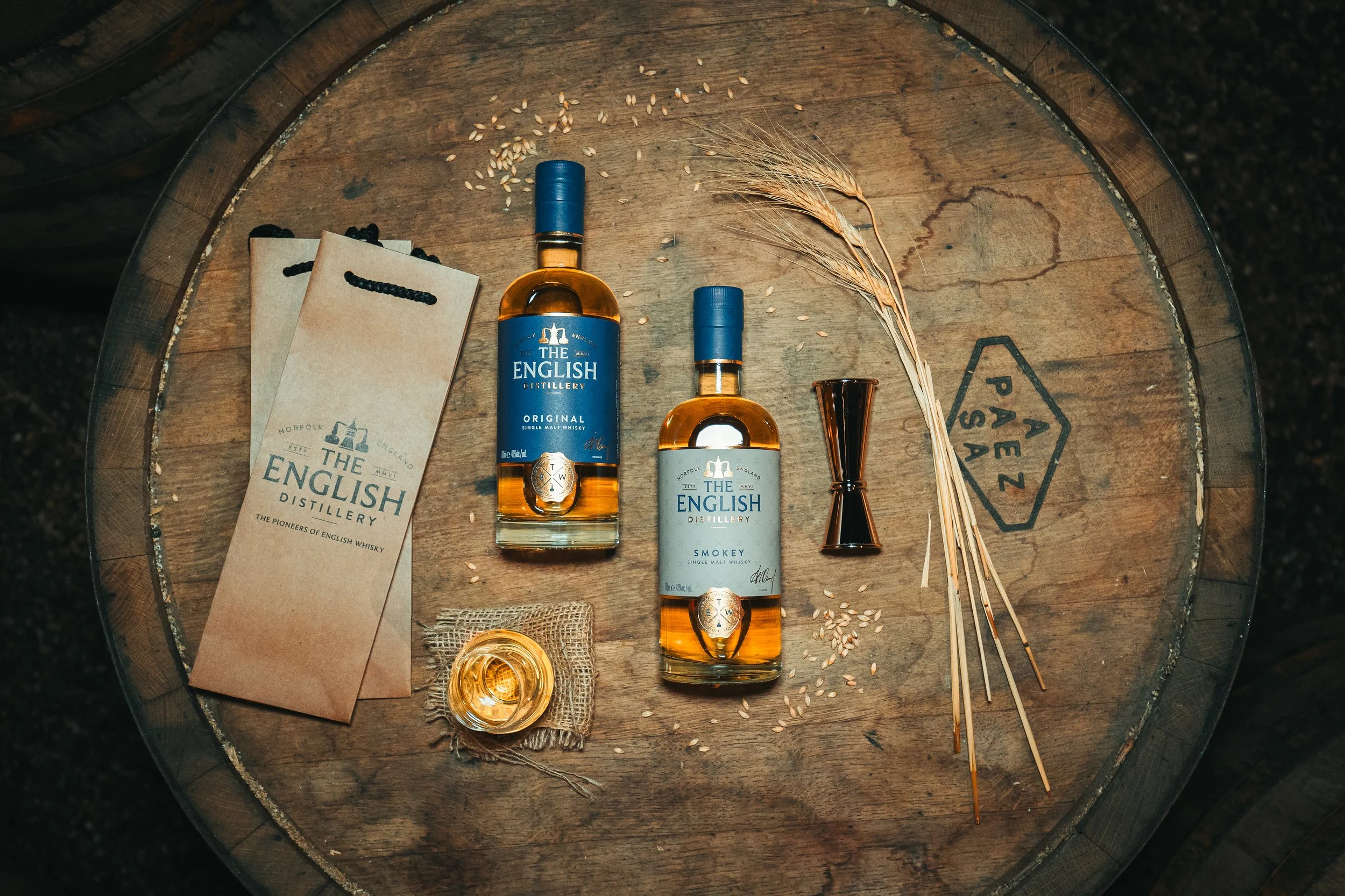 Two bottles of The English Distillery whiskey, one labeled 'Original' and the other 'Smokey,' with a shot glass filled with whiskey, a small burlap cloth, a miniature jigger, some wheat stalks, and scattered rice grains on a wooden barrel surface.