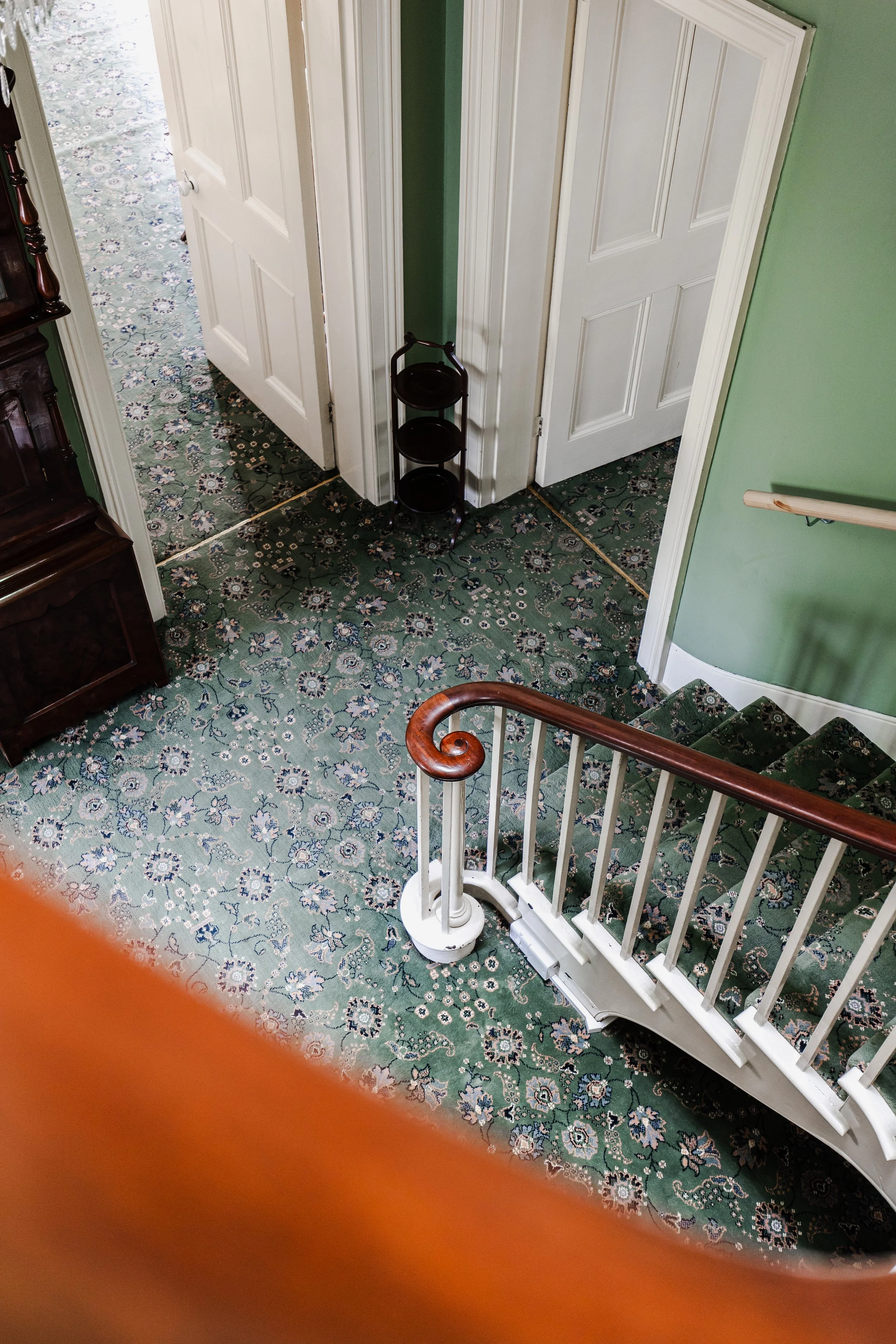 Interior view of a house staircase landing with green walls, a wooden railing, a patterned carpet, and white doors, with a small black three-tier stand near the doorways.
