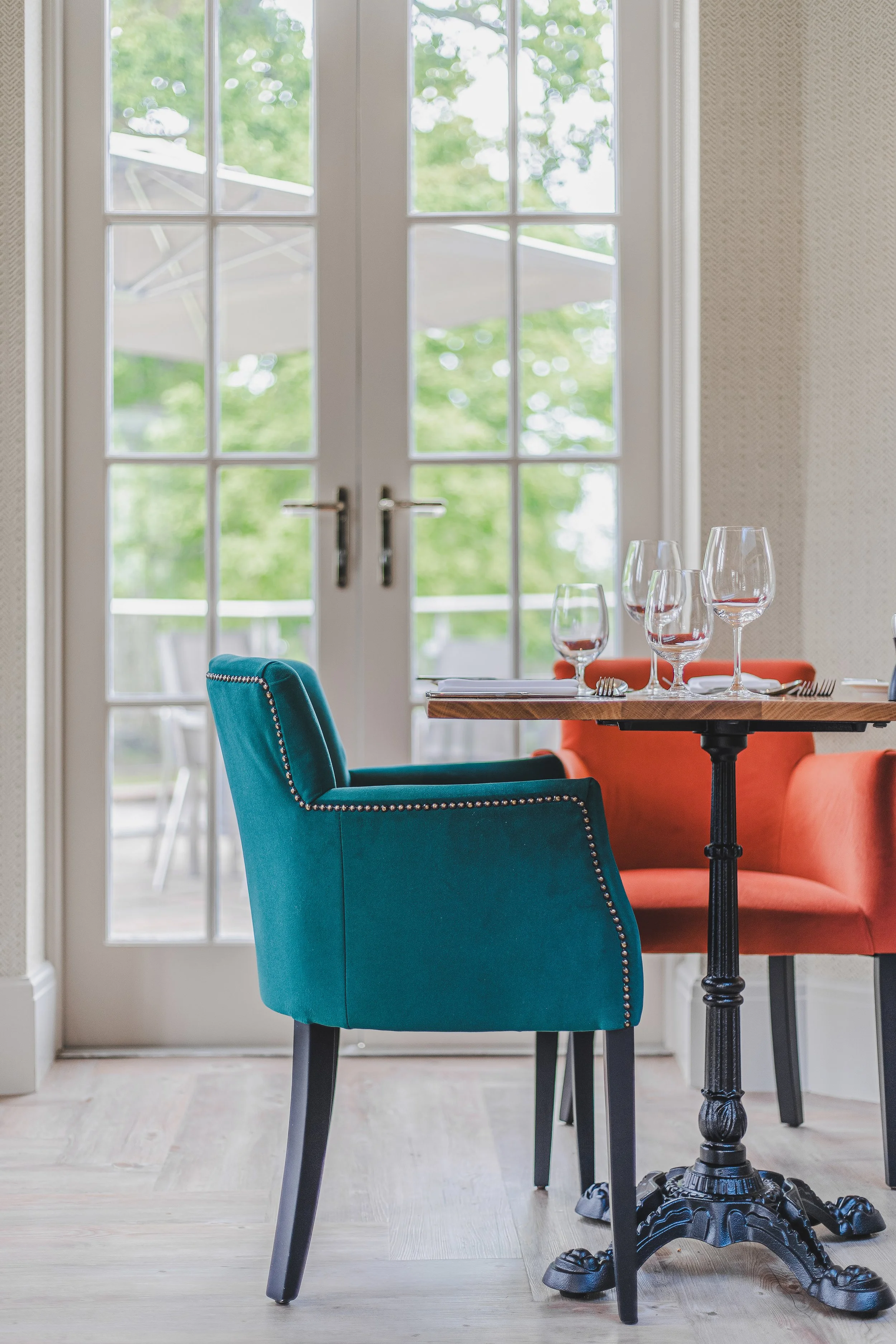 Interior of a dining room with a blue upholstered chair, a wooden table set with wine glasses, and a view through glass doors to an outdoor patio with a large umbrella and green trees.