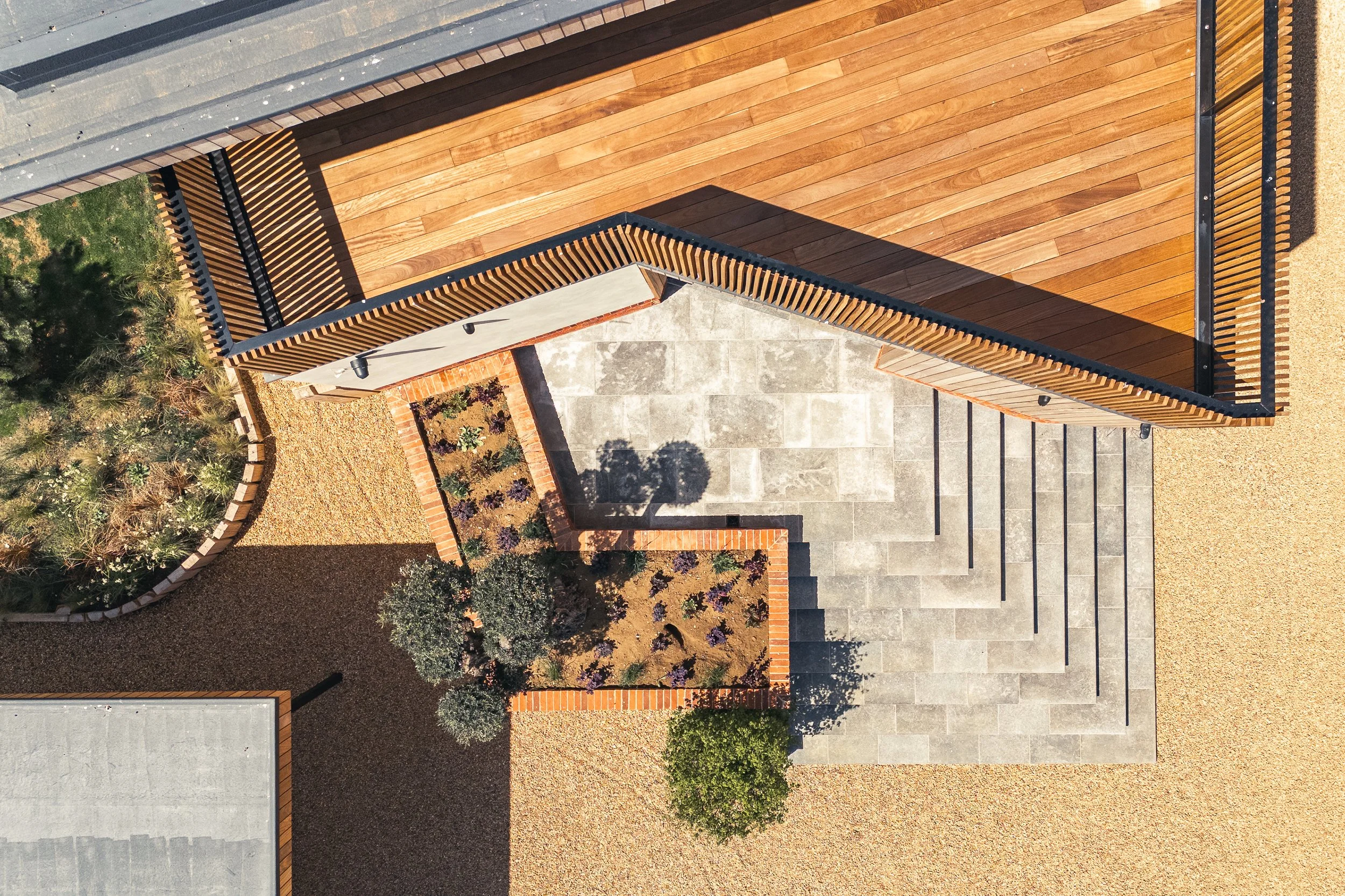 Top-down aerial view of a modern house with wooden and concrete exterior, concrete steps leading to a patio, landscaped garden with small trees and plants, surrounded by gravel and paving stones.