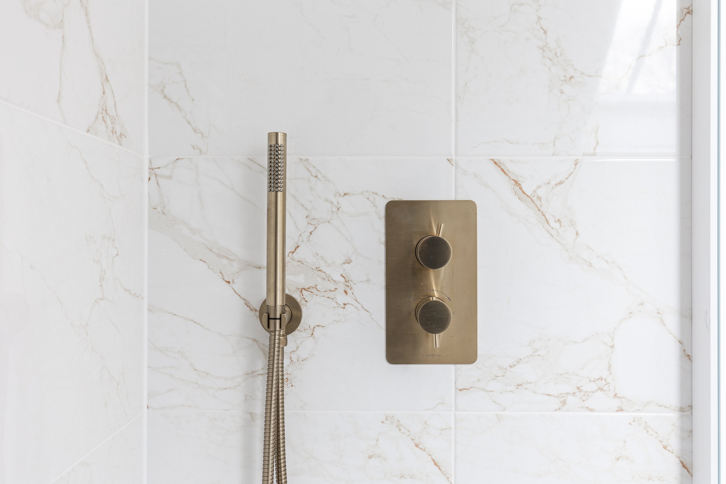 Close-up of a wall-mounted shower panel with a handheld showerhead, two round control knobs, and a marble tile background.