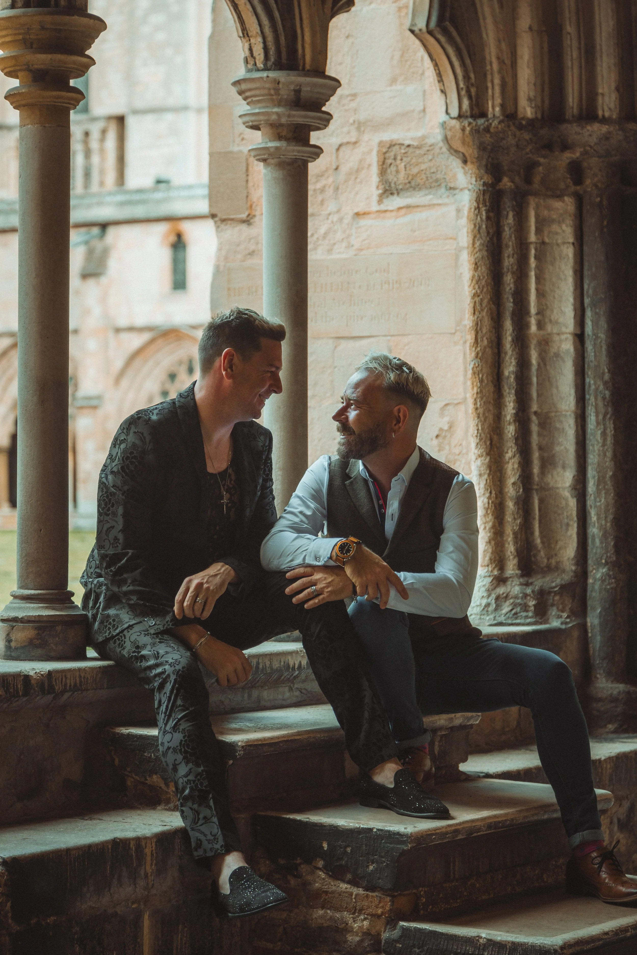 Two men sitting on the steps of an ancient stone building, smiling and engaged in conversation, with medieval-style arches in the background.