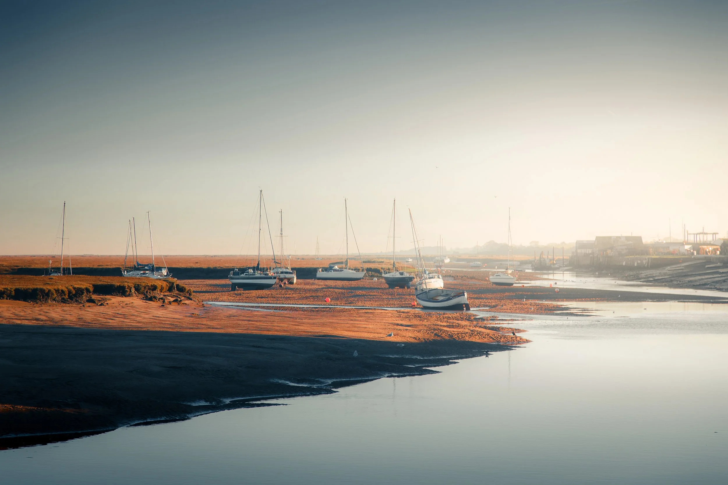 A quiet harbor with sailboats on the shore during sunset or early morning, reflecting calm waters with a few buildings in the background.
