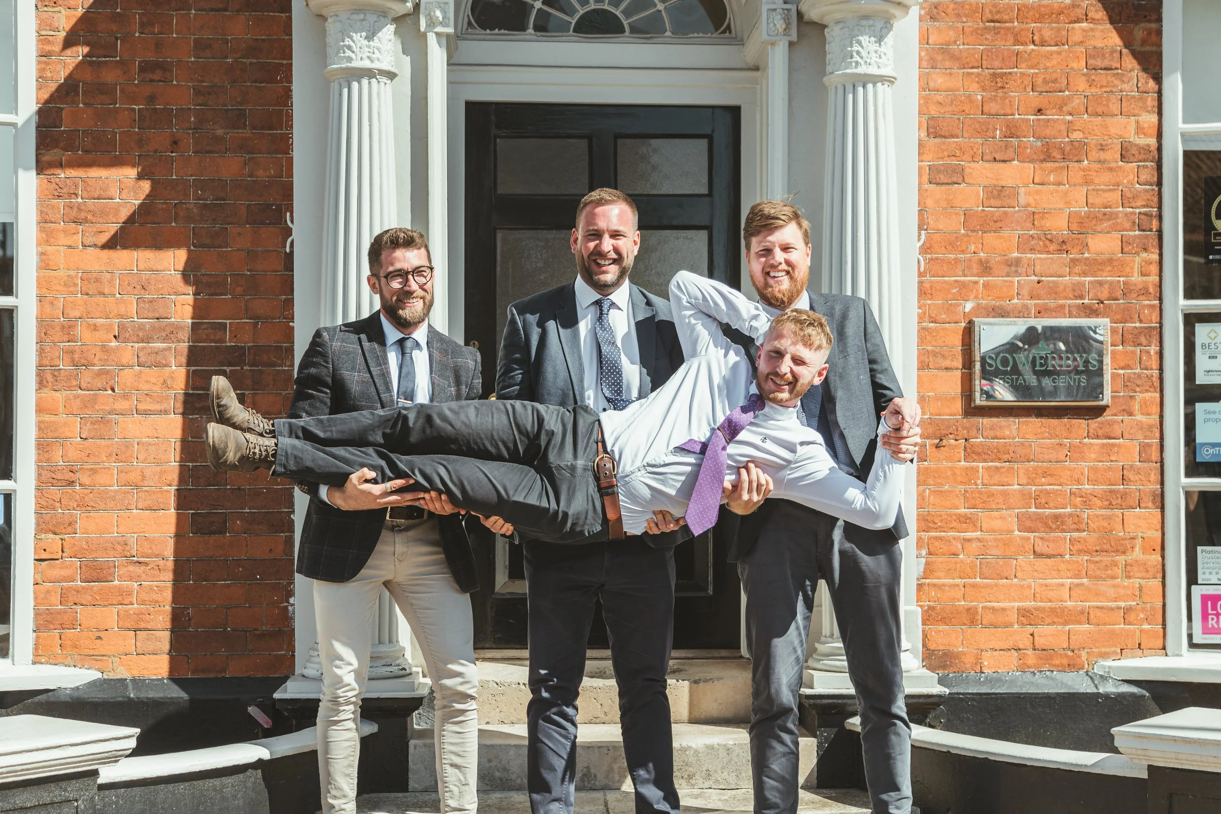 Five men in suits posing in front of a brick building. One man is being held horizontally by the other four men, all smiling and dressed in business attire.