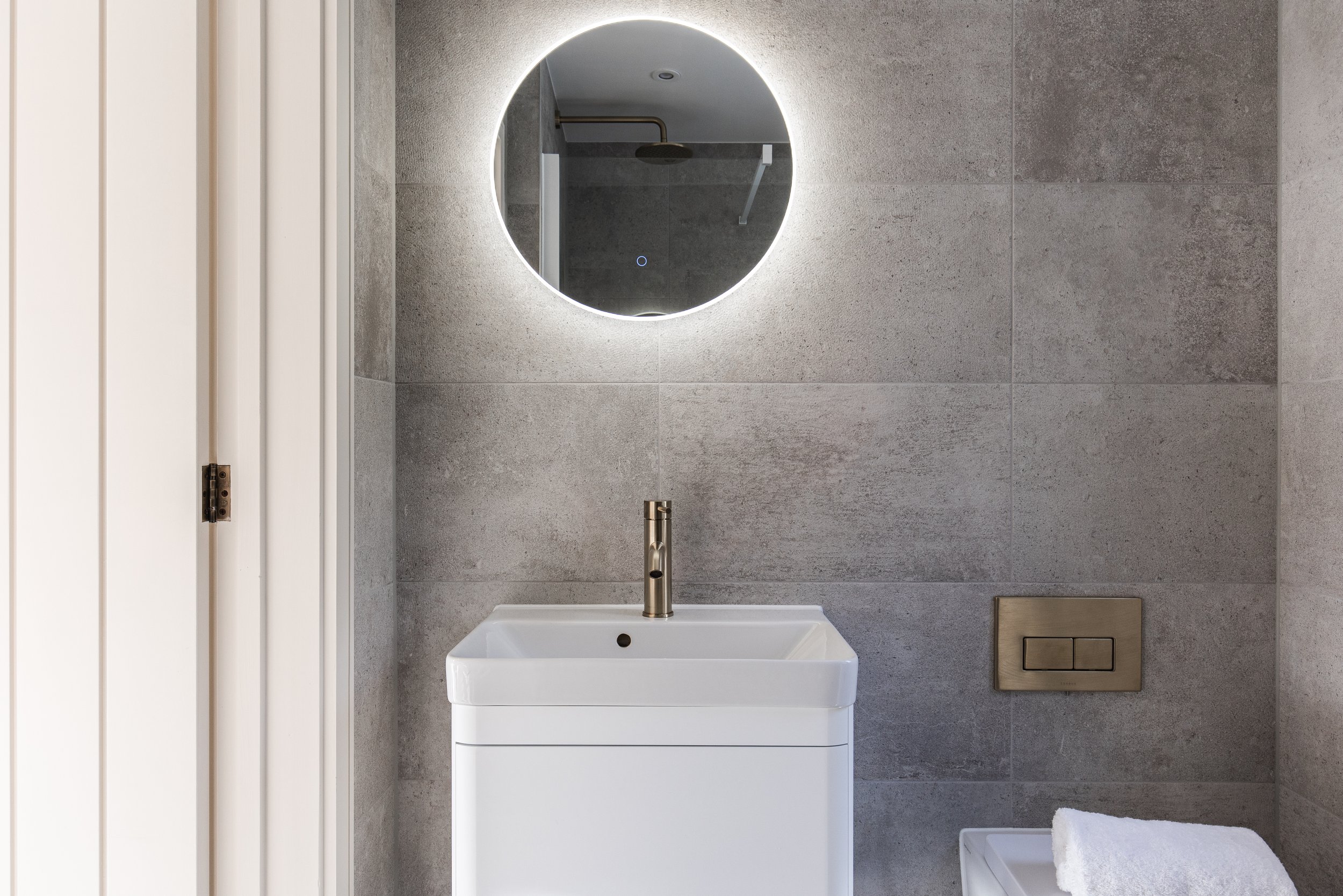 Modern bathroom with a round illuminated mirror, a white sink with a silver faucet, a wall-mounted flush button, and a rolled towel on a counter.