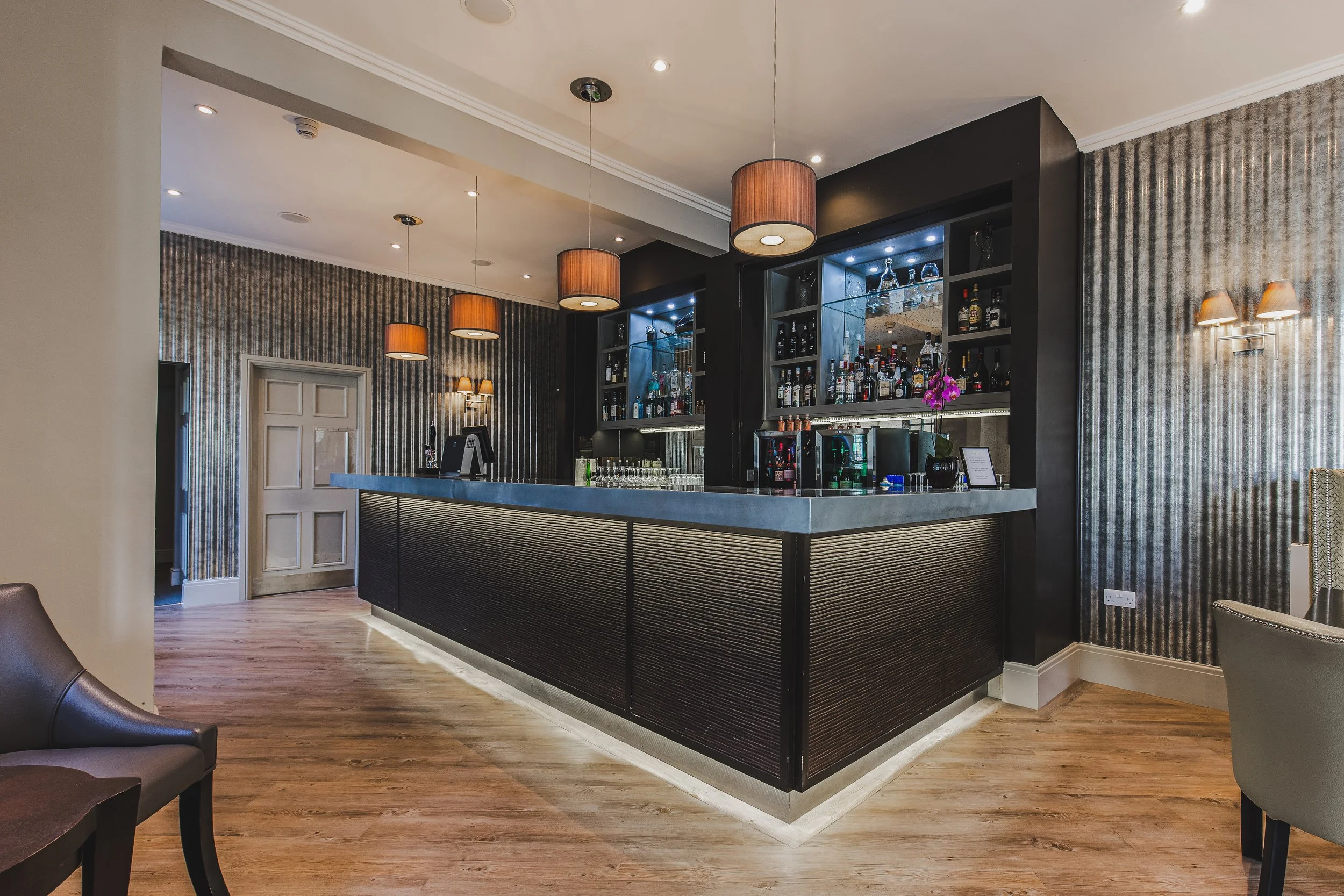 A modern hotel bar with a sleek black finish, featuring illuminated shelves stocked with liquor bottles and glassware, decorated with a purple orchid, and surrounded by patterned wallpaper and wooden floors.